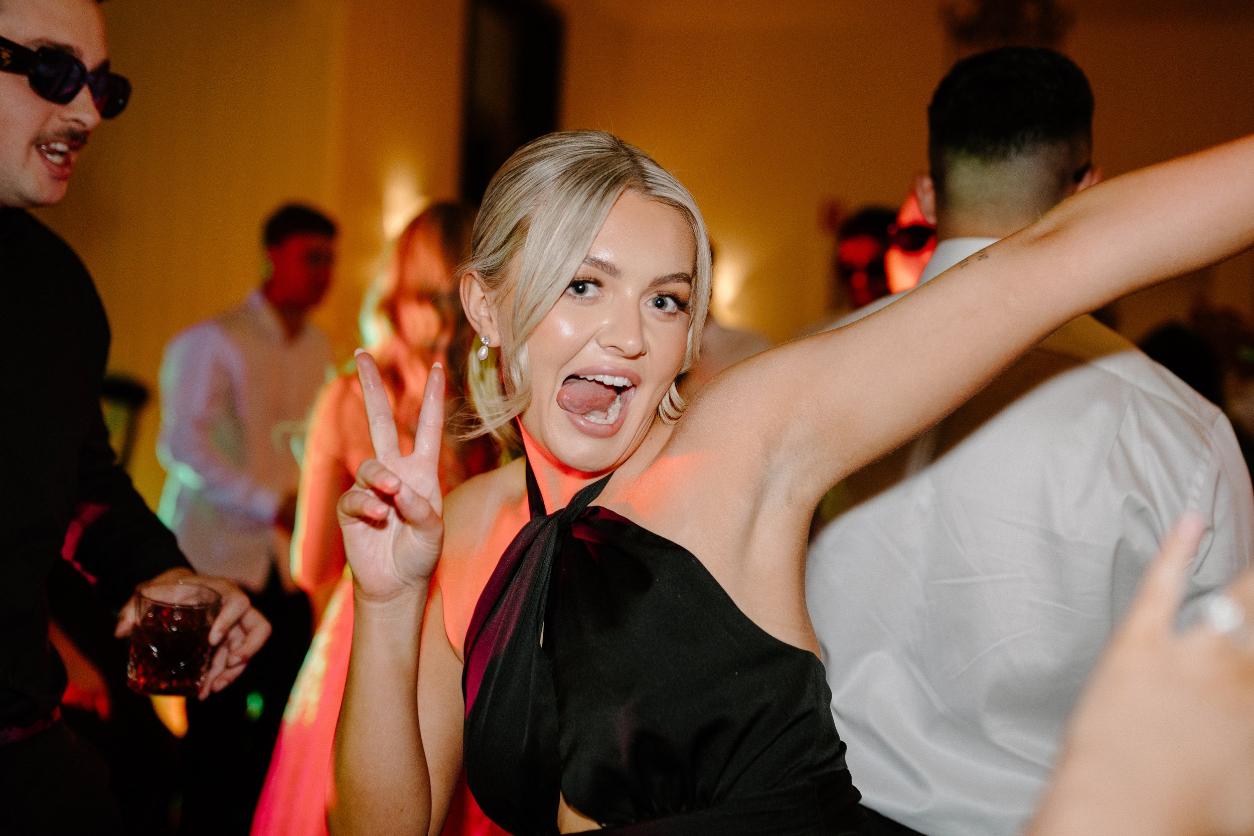 A young woman with blonde hair making a peace sign and posing playfully at a lively party or dance event, with other people dancing and socializing in the background.