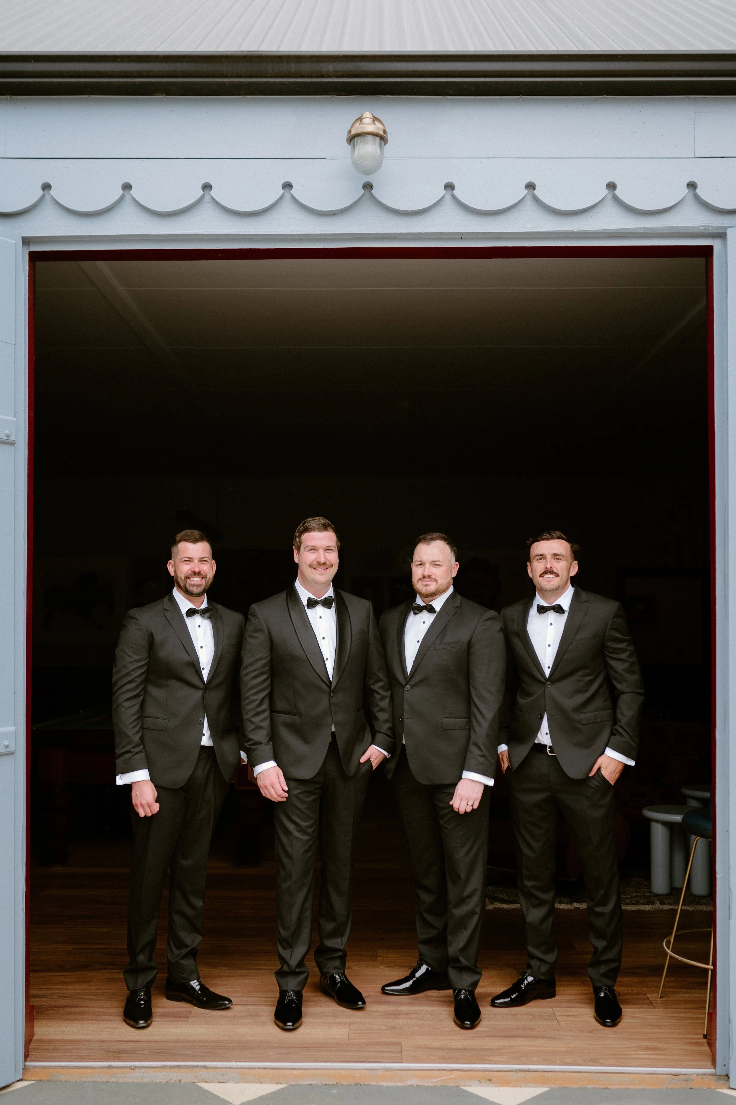 Four men in tuxedos standing inside a garage, smiling for a photo.