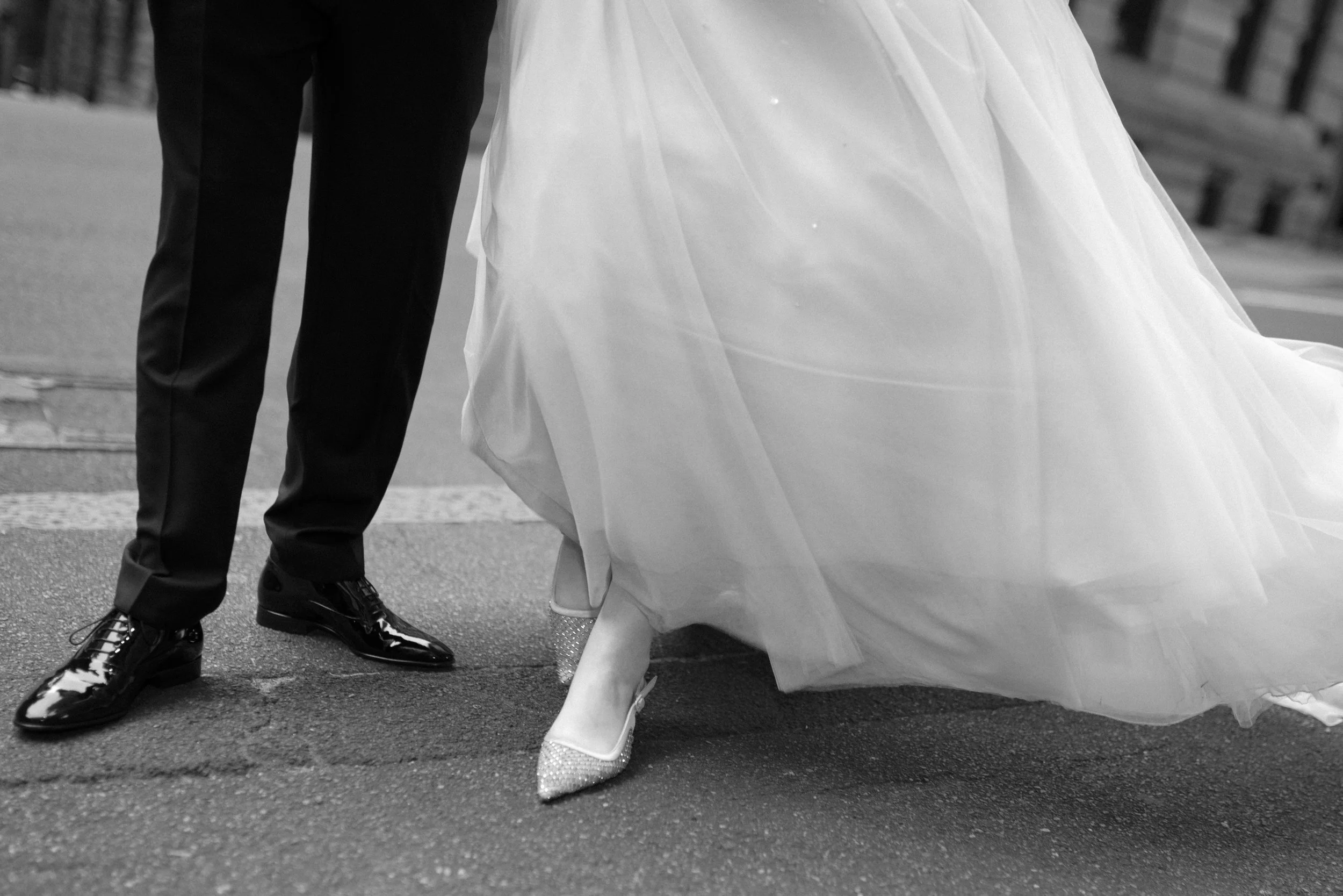 Close-up of a couple's legs and feet, with one woman in sparkling heels and a flowing wedding dress and a man in polished dress shoes and suit pants, walking on a street.