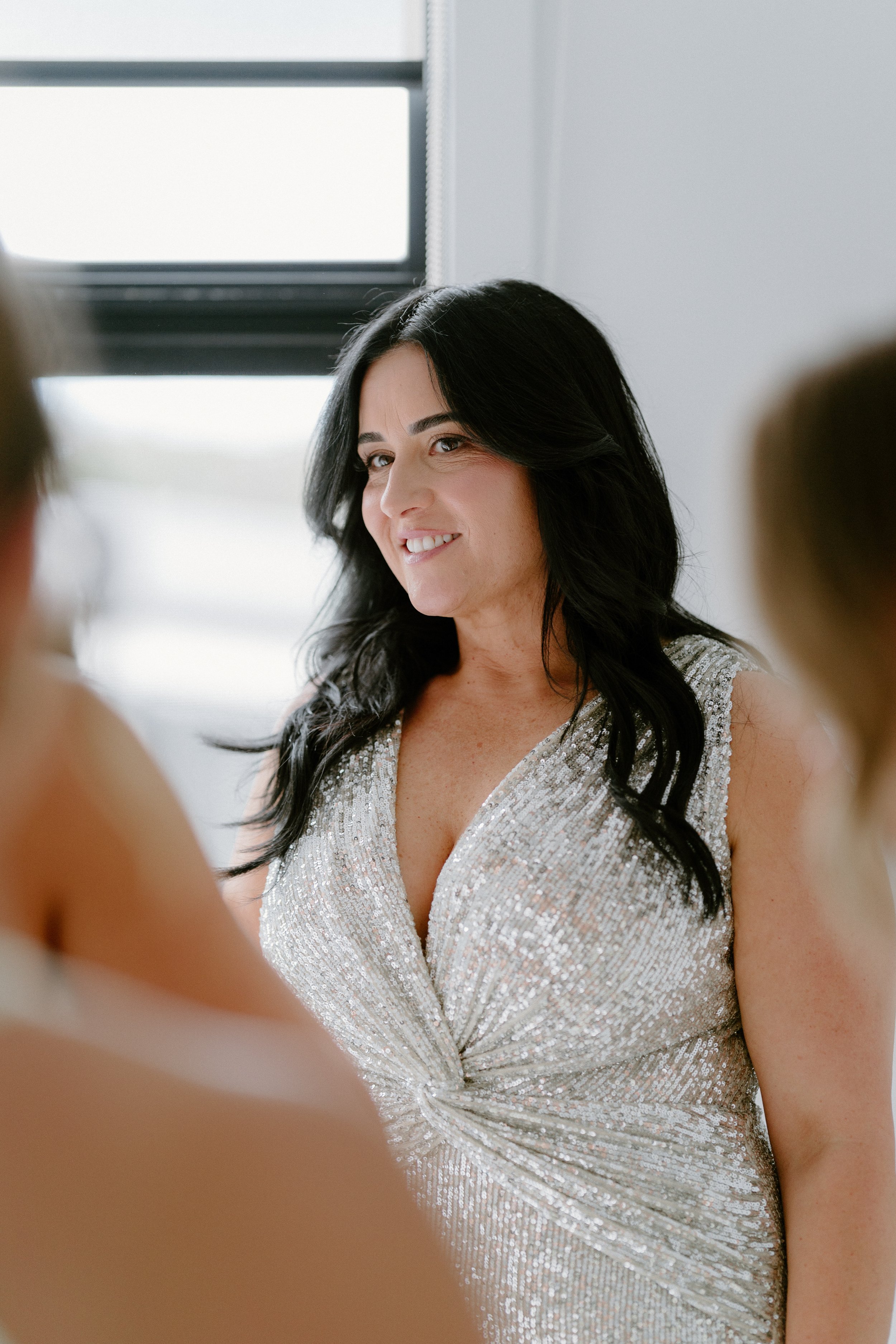 A woman with long black hair in a sparkly silver dress, smiling and looking to the side, indoors near a window.