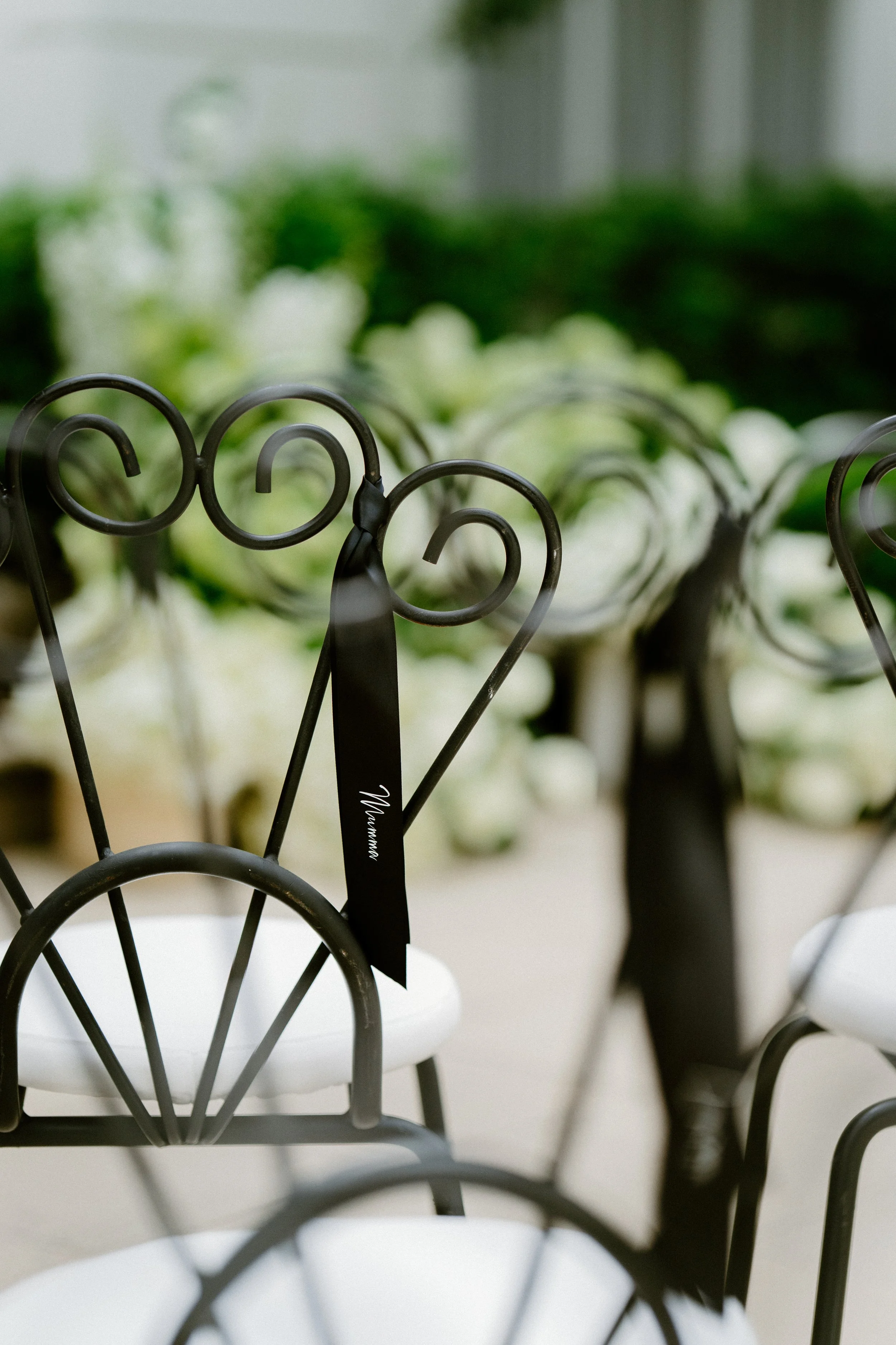 Close-up of a black wrought iron chair with a white cushion, decorated with a black ribbon with white text, outside with blurred white flowers and greenery in the background.