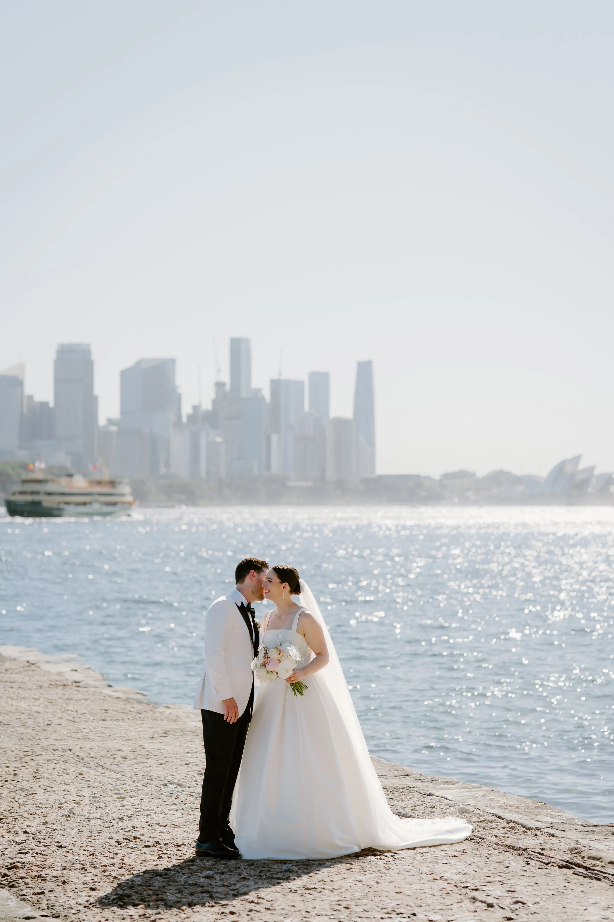 Sergeants Mess, Mosman Wedding Venue. Sergeants Mess Wedding Photos. A bride and groom in wedding attire standing on a rocky waterfront, sharing a kiss with the city skyline and Sydney Opera House in the background.