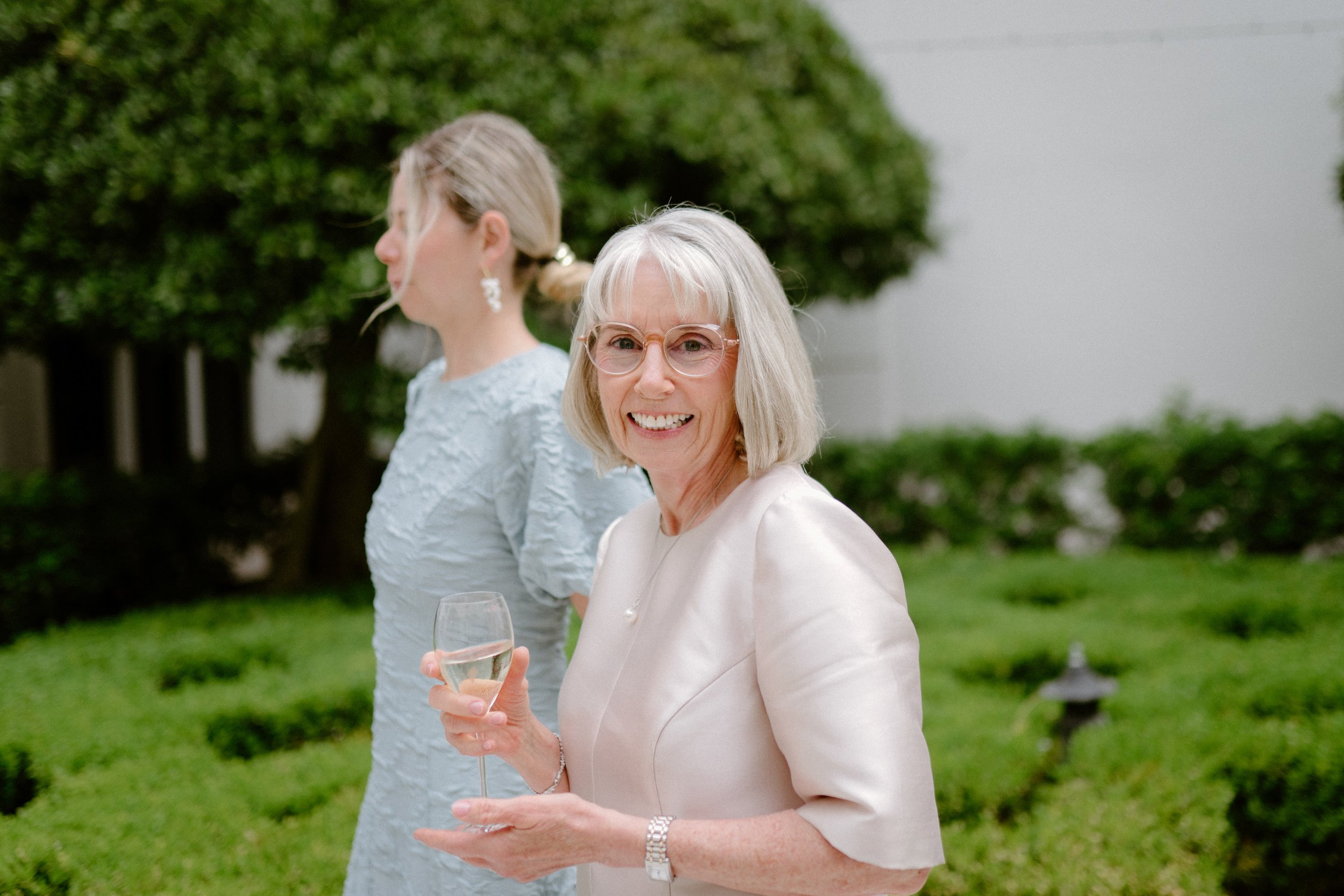 A smiling older woman with short gray hair and glasses holding a glass of white wine, standing outside in a garden with green trees and bushes. In the background, a younger woman with blonde hair in an updo wearing a light blue dress is turned sidewa