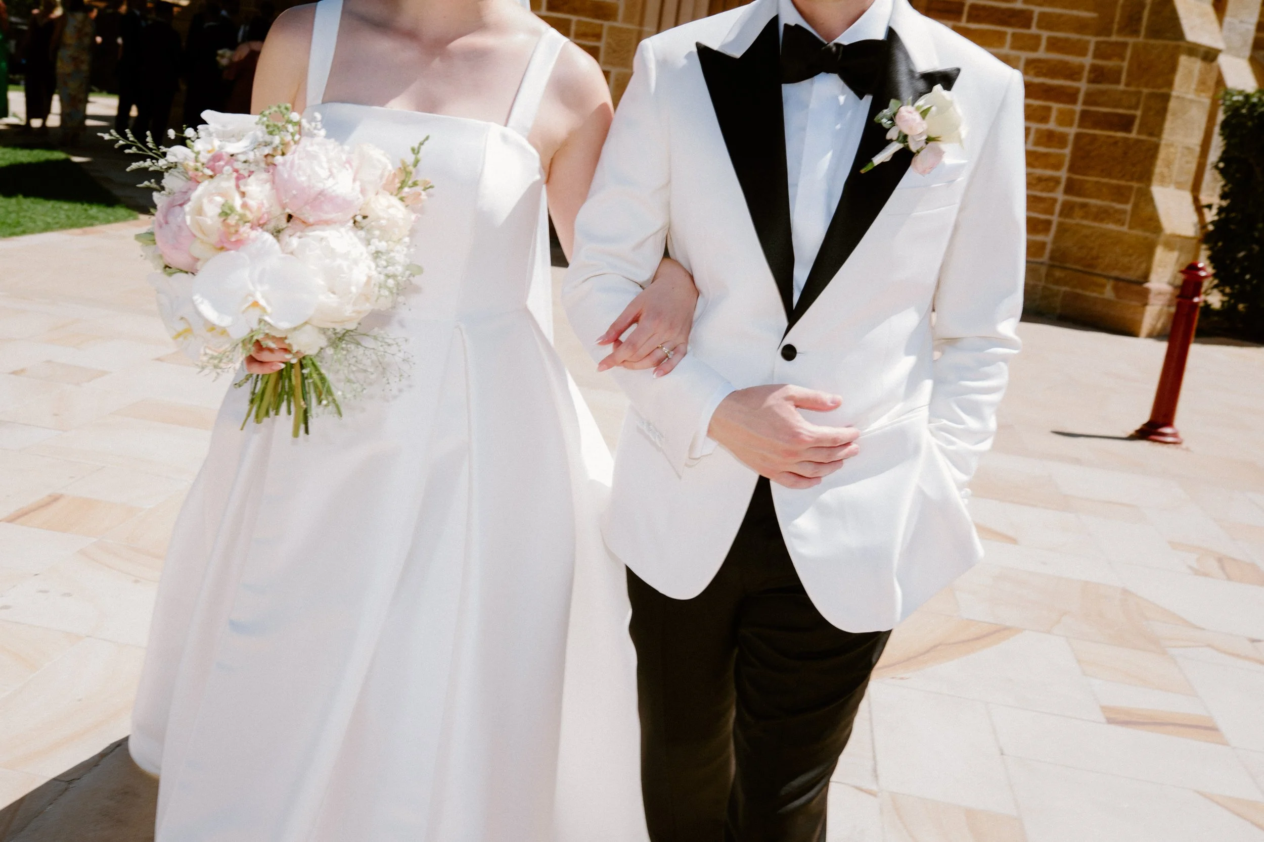 Bride and groom walking outside during wedding, bride holding bouquet, groom in tuxedo with black bowtie, brick building in background.