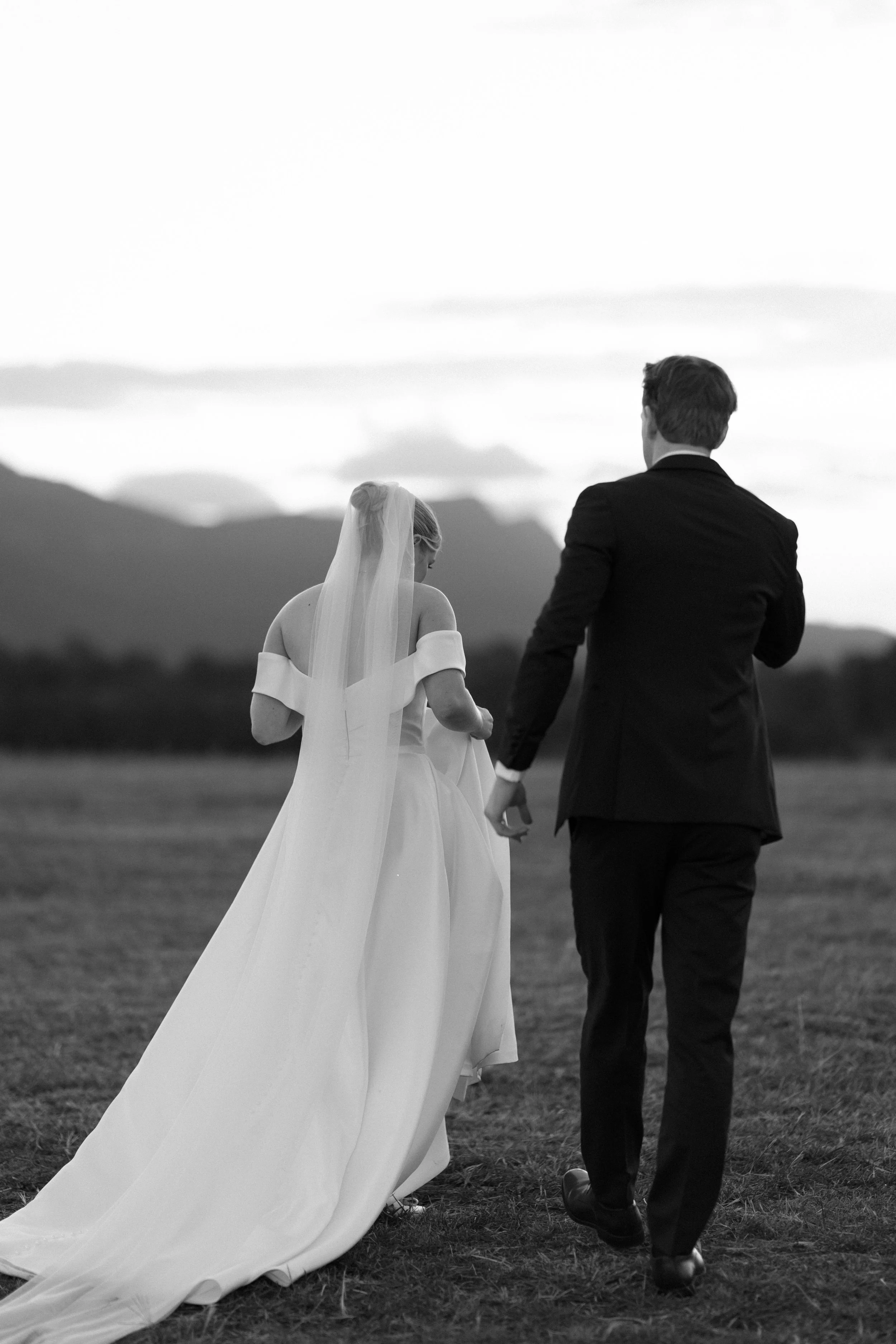 A black-and-white photo of a bride and groom walking away together outdoors, with mountains and a cloudy sky in the background. The bride is wearing a wedding dress and veil, and the groom is in a dark suit.