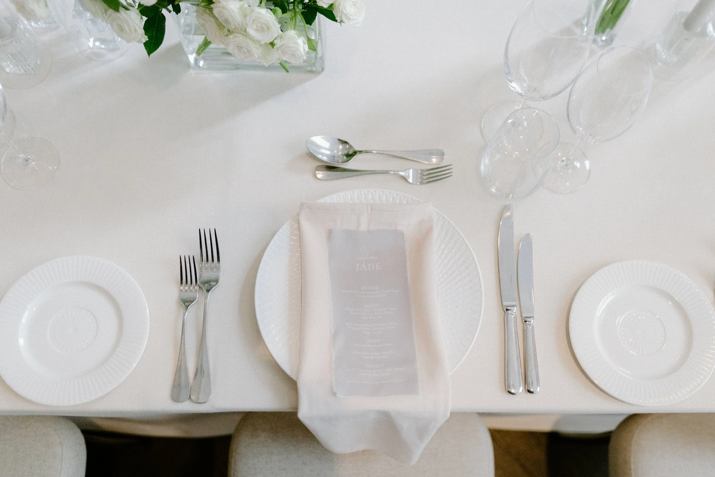 Elegant place setting with white plates, silverware, stem glasses, and a flower arrangement in a glass vase on a white tablecloth.