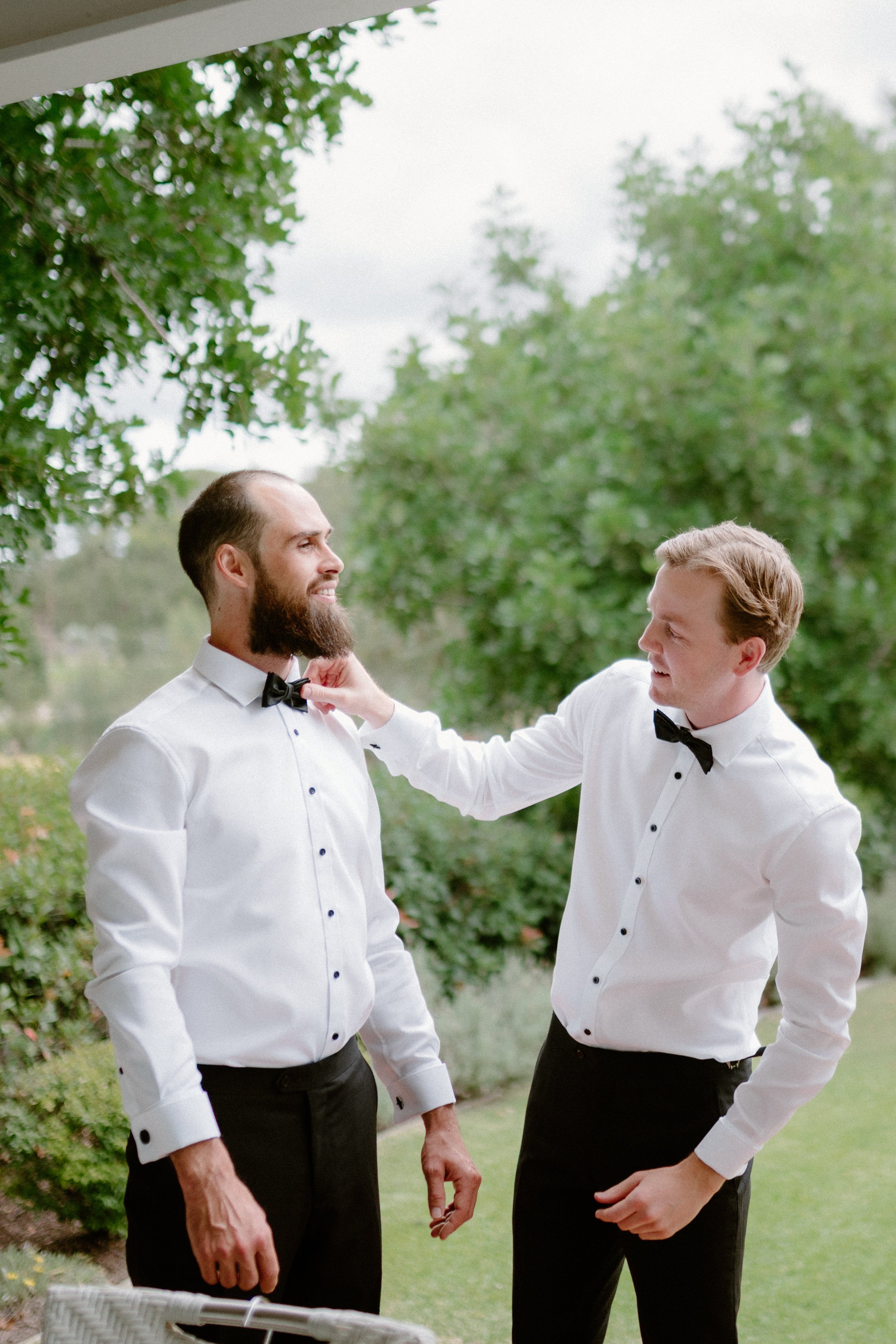 Two men dressed in white tuxedos with black bow ties standing outdoors in a lush green garden. One man is adjusting the bow tie of the other, both smiling.
