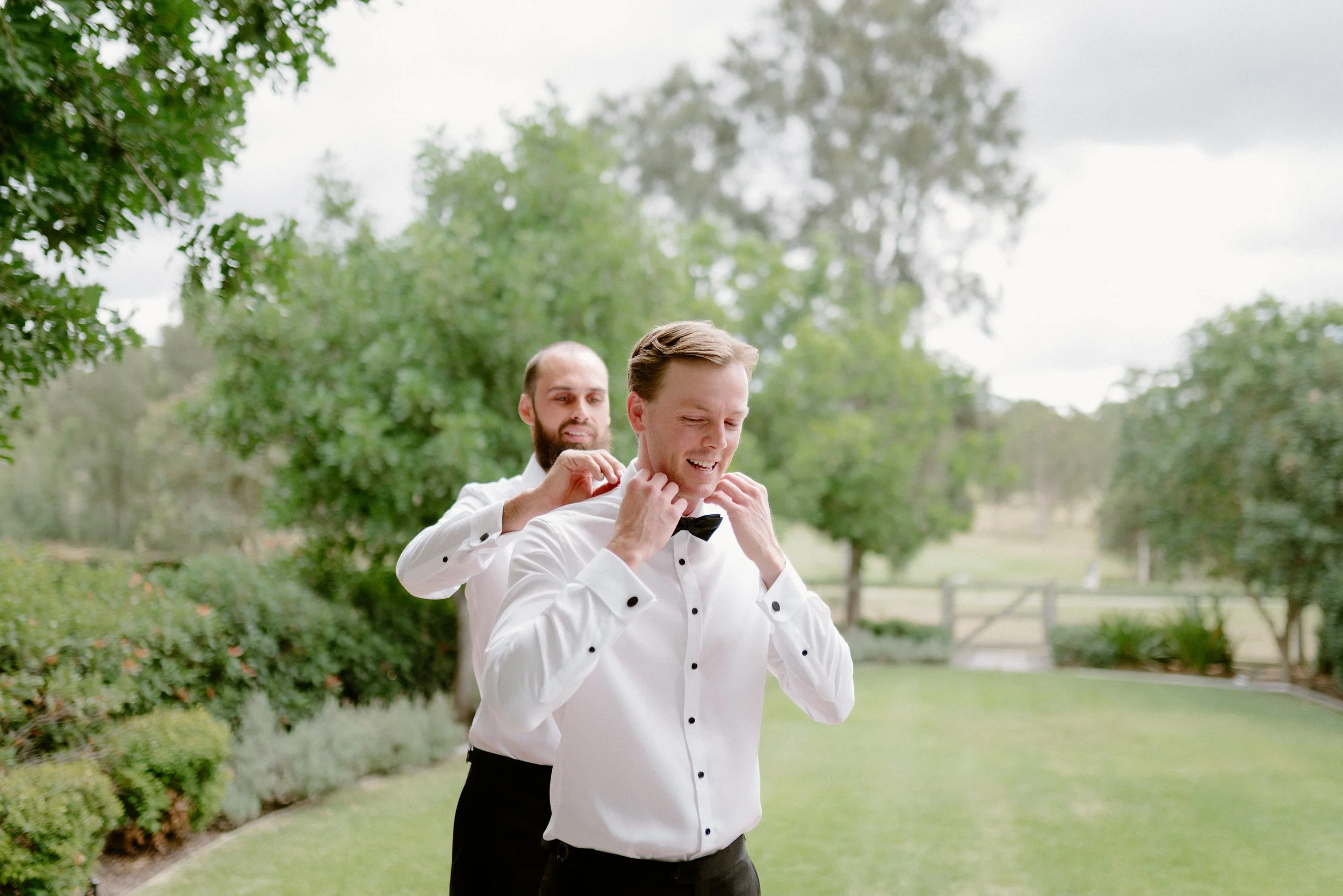 Two men in tuxedos standing outdoors in a park-like setting with green trees and grass, one helping the other adjust his bow tie.