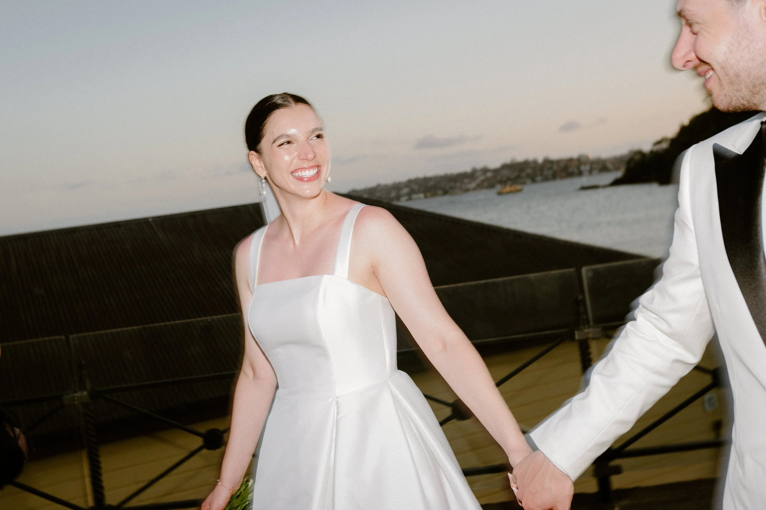 A newlywed couple is holding hands and smiling at each other outdoors during sunset, with water and a distant shoreline in the background.