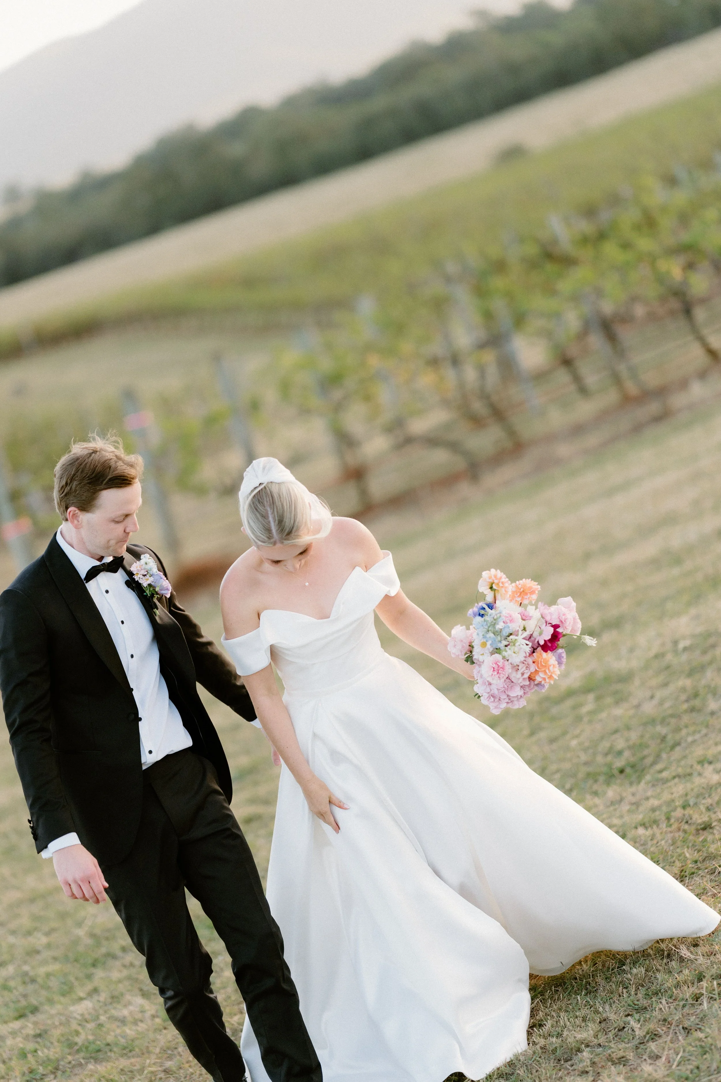 A bride and groom walking outdoors during a wedding ceremony, with a vineyard in the background. The bride is wearing a white off-the-shoulder dress and holding a bouquet of pink, purple, and white flowers. The groom is wearing a black tuxedo with a 