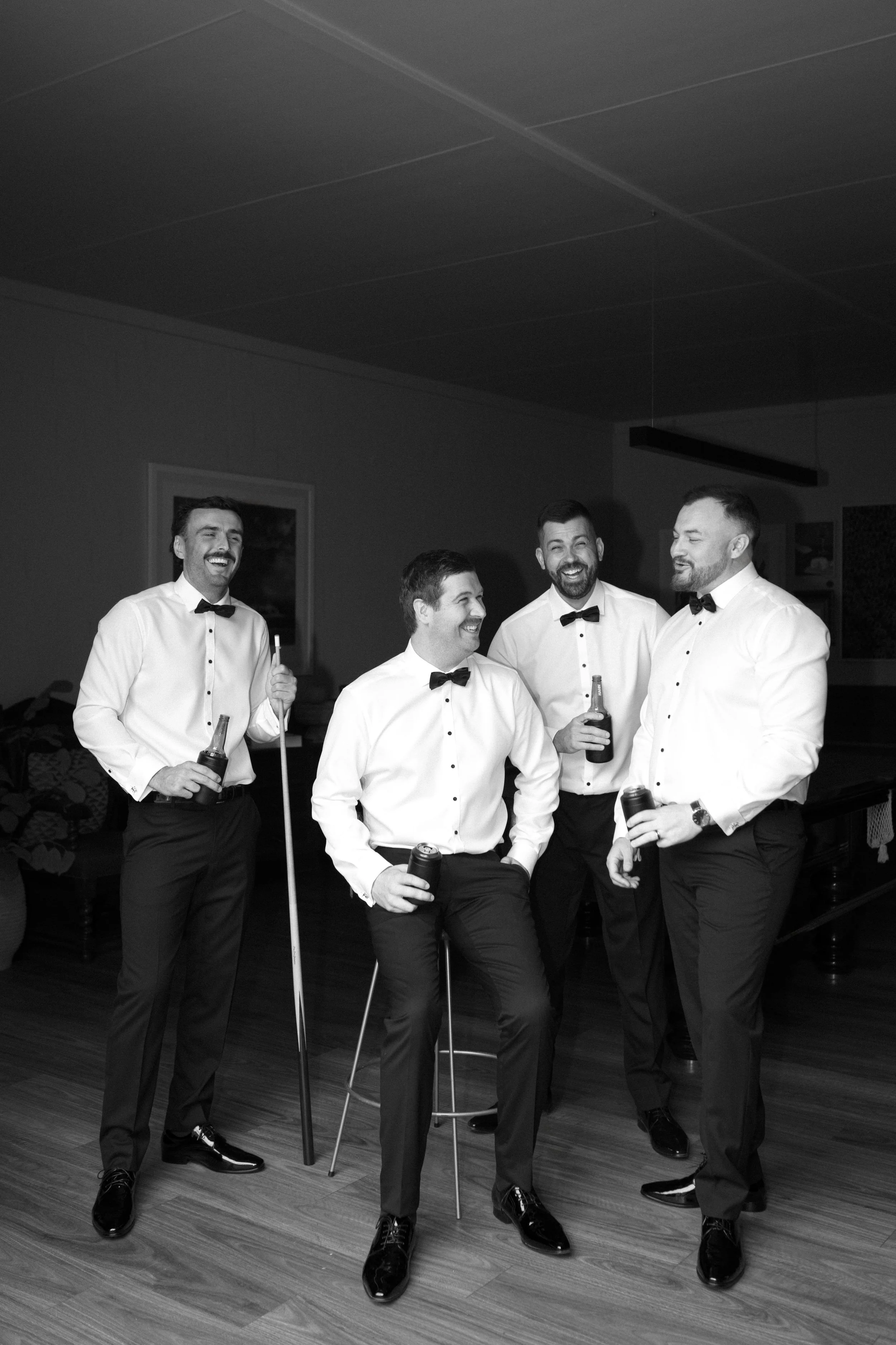 The Sienna, Hunter Valley Wedding Venue, Wedding Photos. Black and white photo of four men in tuxedos with bow ties, smiling and holding drinks, standing and sitting in a room with wooden floors, celebrating or socializing.