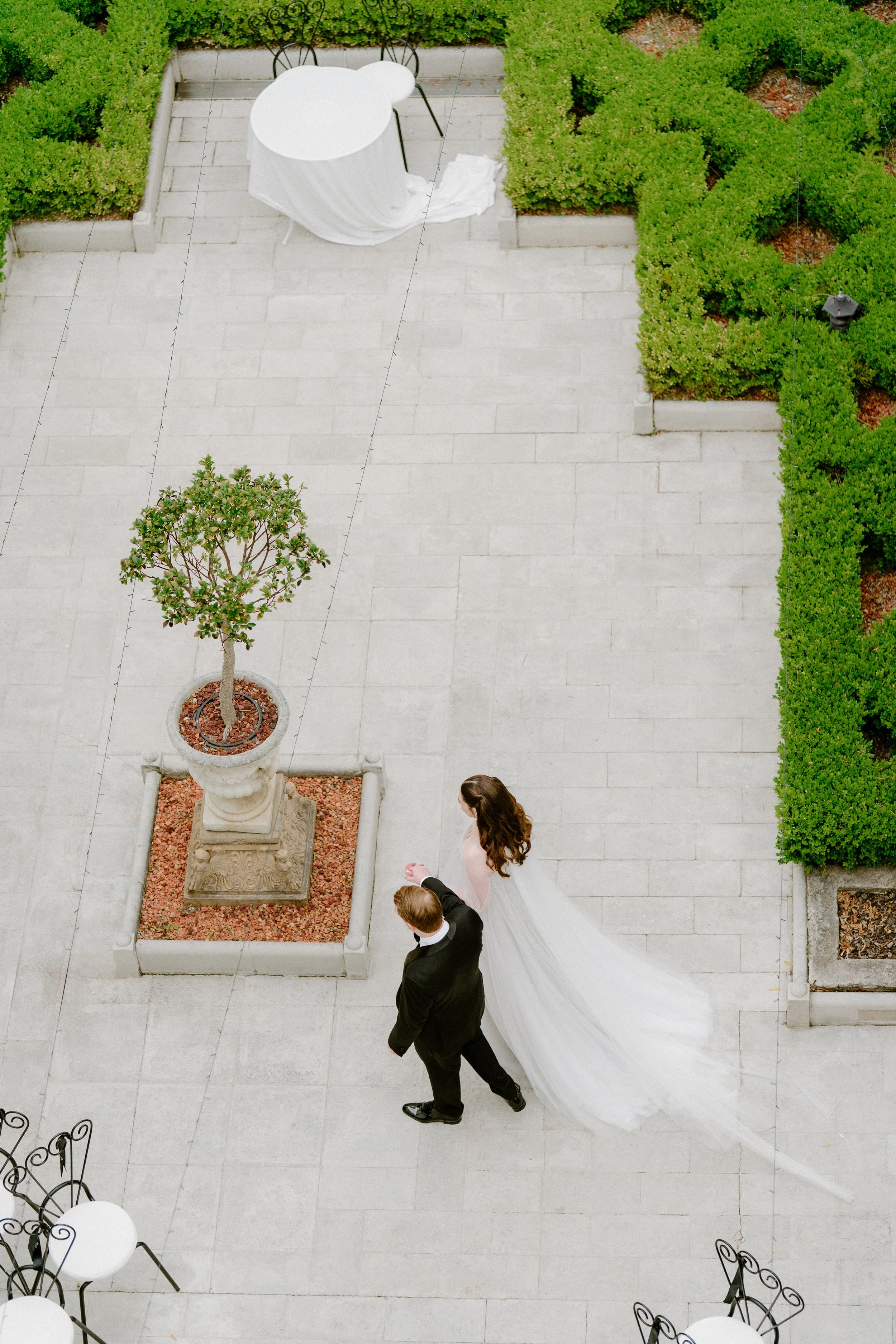 A bride in a white wedding dress and a groom in a black suit walking hand in hand in an outdoor garden area with stone tile flooring, surrounded by green hedges and a small tree in a planter.