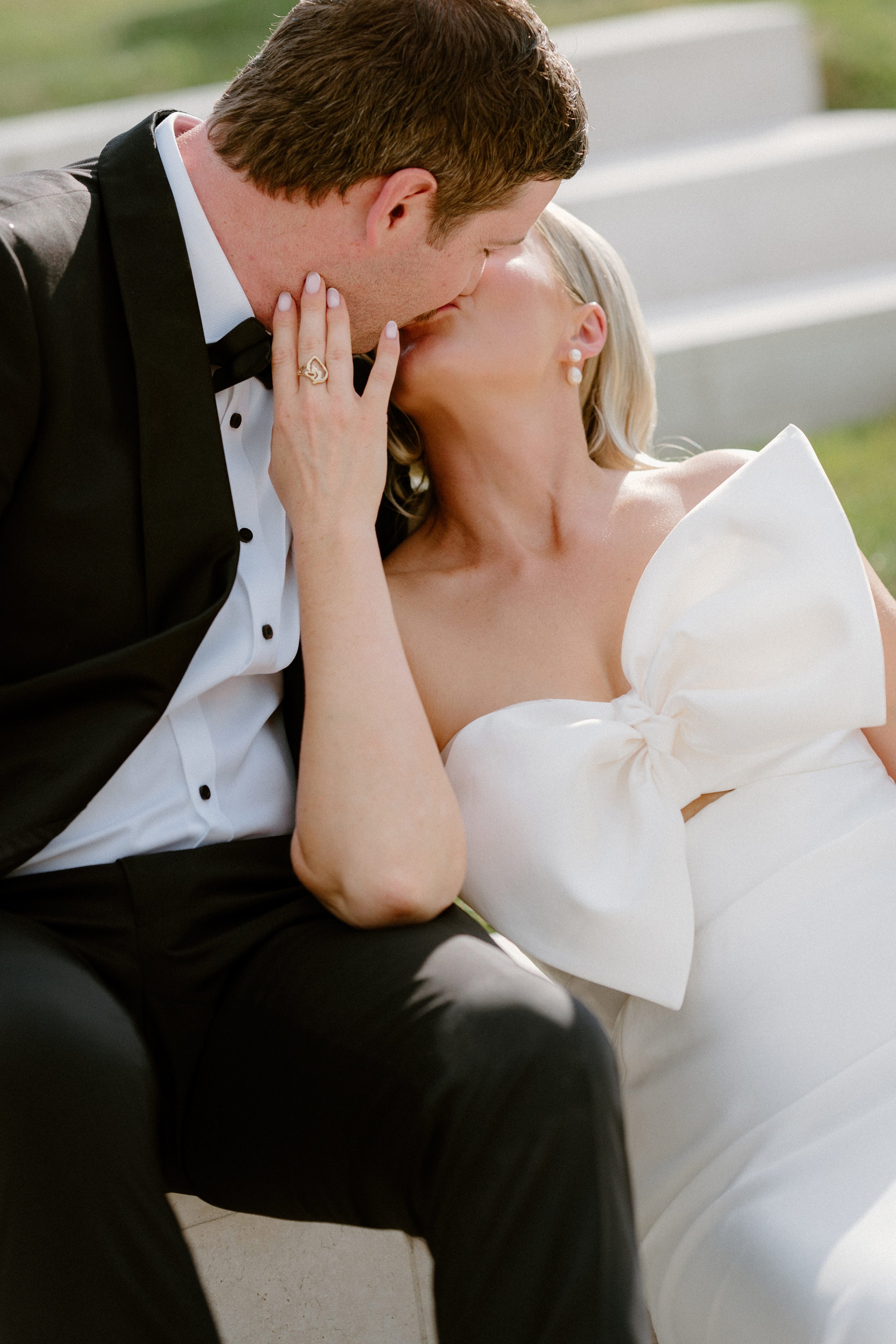 A bride and groom sharing a kiss, with the bride wearing a white dress with a large bow and pearls, and the groom in a tuxedo, outdoors on a sunny day.