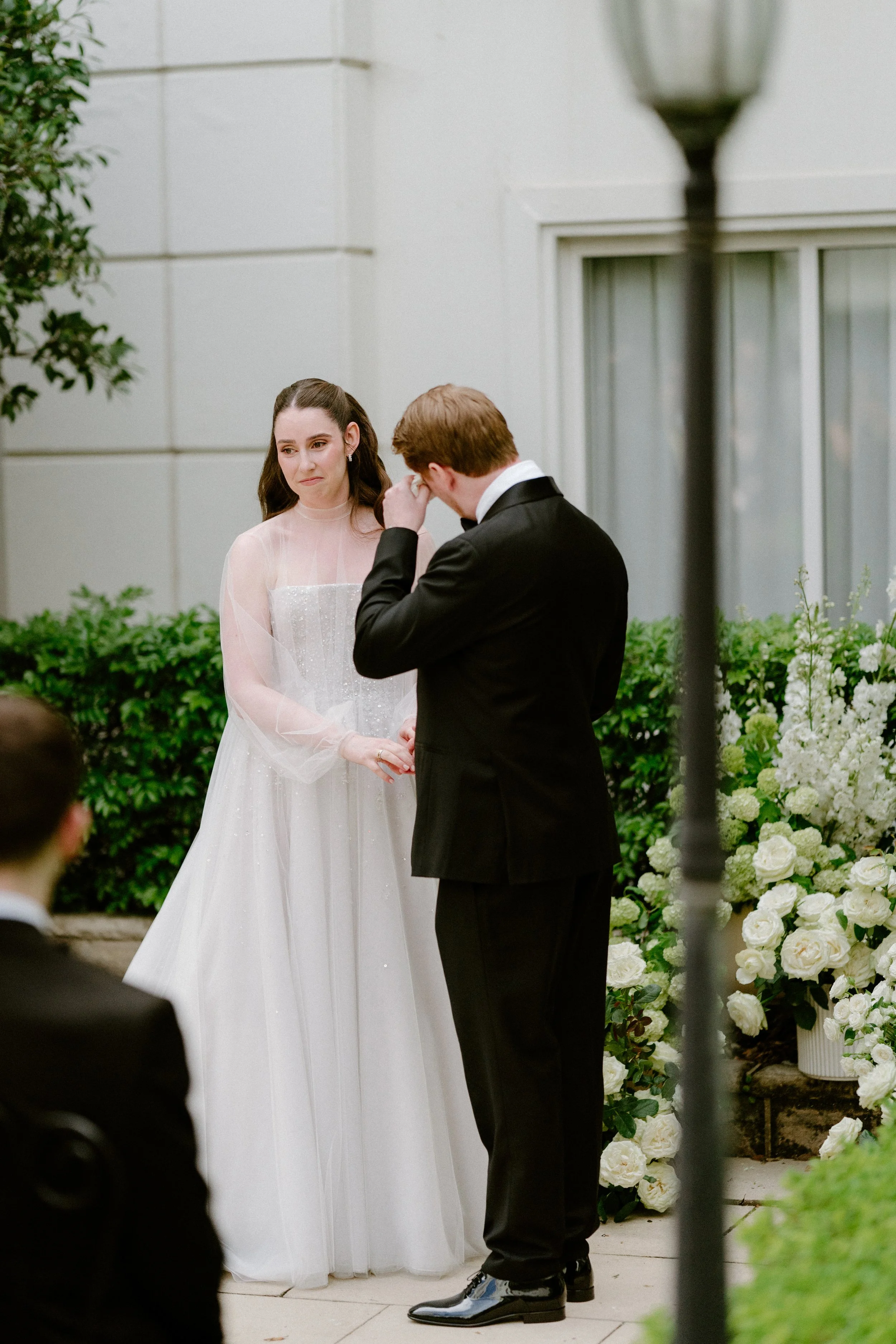 A wedding ceremony with a bride in a white gown and a groom in a black tuxedo, standing outdoors near white floral arrangements.