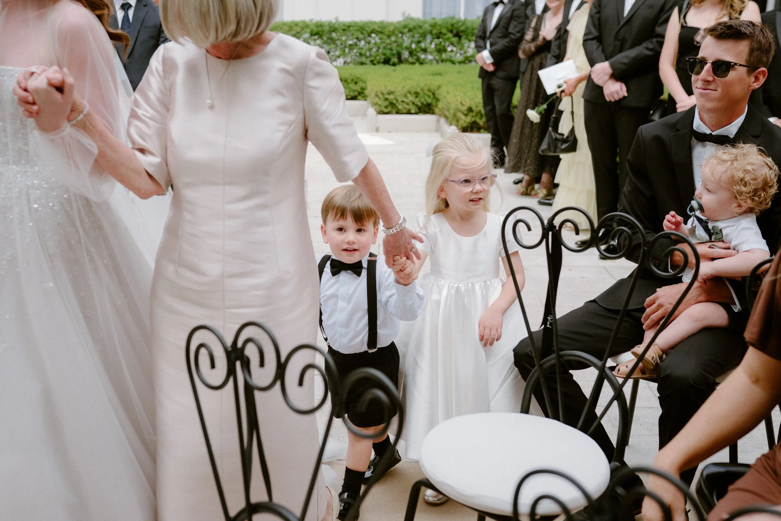 Children, including a boy in a white shirt with a black bow tie, a girl with glasses in a white dress, and a young boy with curly hair in a tuxedo, are holding hands during a wedding ceremony outdoors, with adults in formal attire standing in the bac