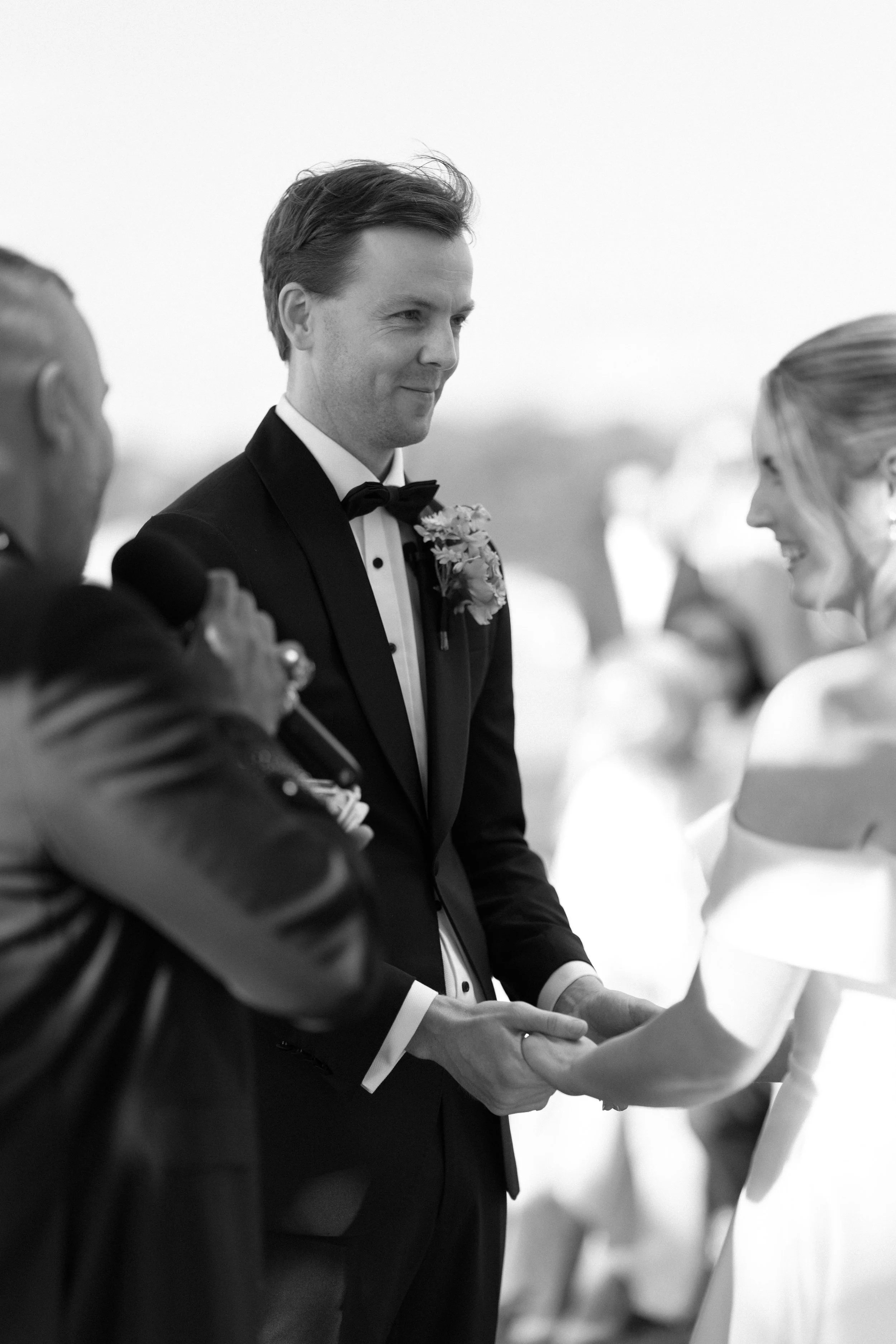 Black and white photo of a groom and bride holding hands during their wedding ceremony, with an officiant speaking to them.