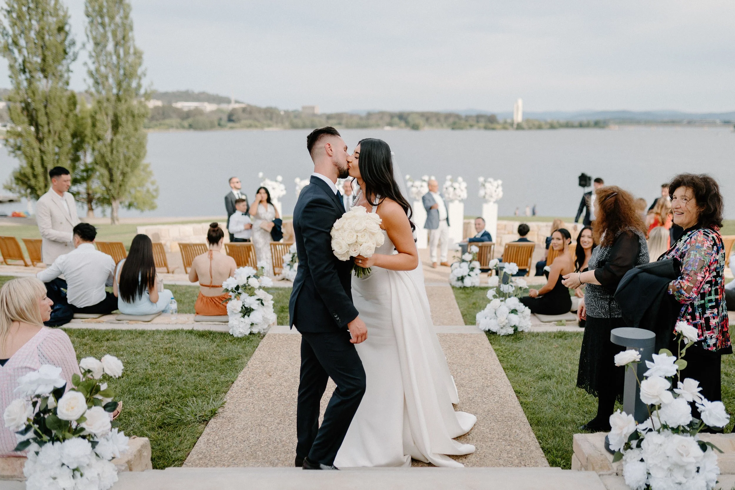 A bride and groom kiss during their outdoor wedding ceremony by a lake, with guests seated on either side, floral decorations, and scenic water and trees in the background.