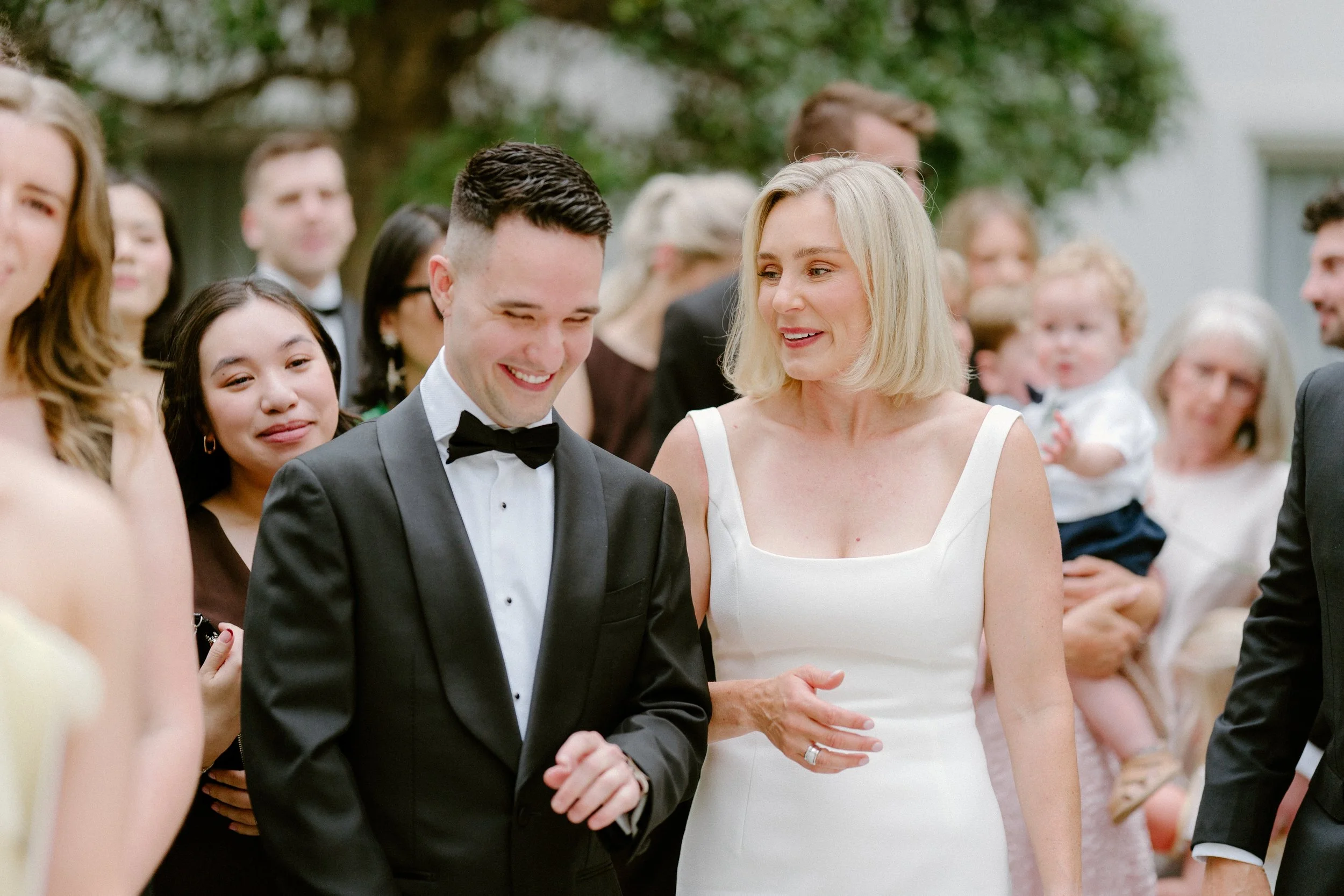 A wedding scene with a smiling groom in a tuxedo and a woman in a white dress, surrounded by guests, outdoors with trees in the background.