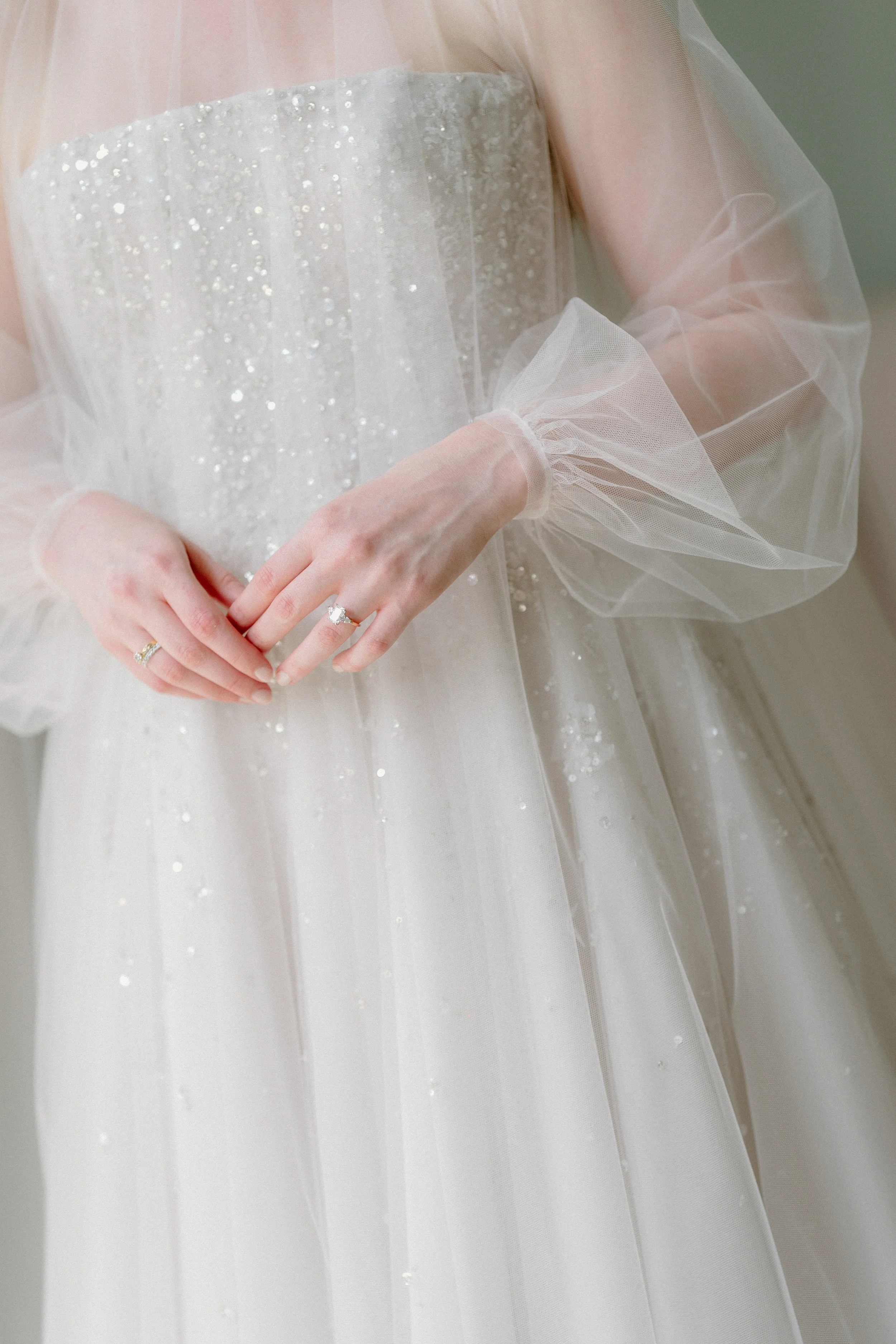 Close-up of a bride wearing a wedding dress with sheer, puffy sleeves and shimmering embellishments, with hands clasped in front.
