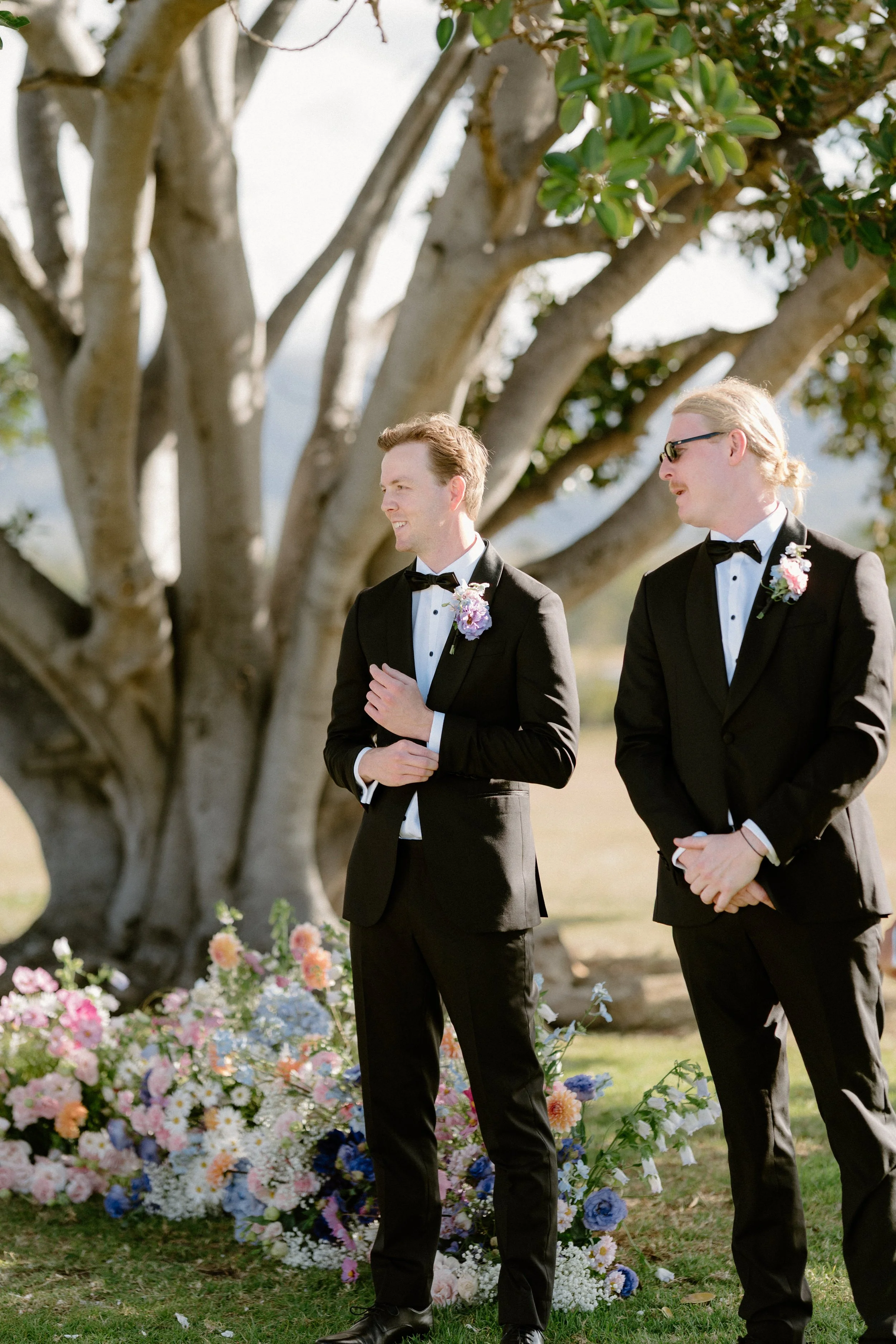 Two men in tuxedos with boutonnières, standing outdoors near a large tree with flowering plants and arranging flowers in a wedding setting.