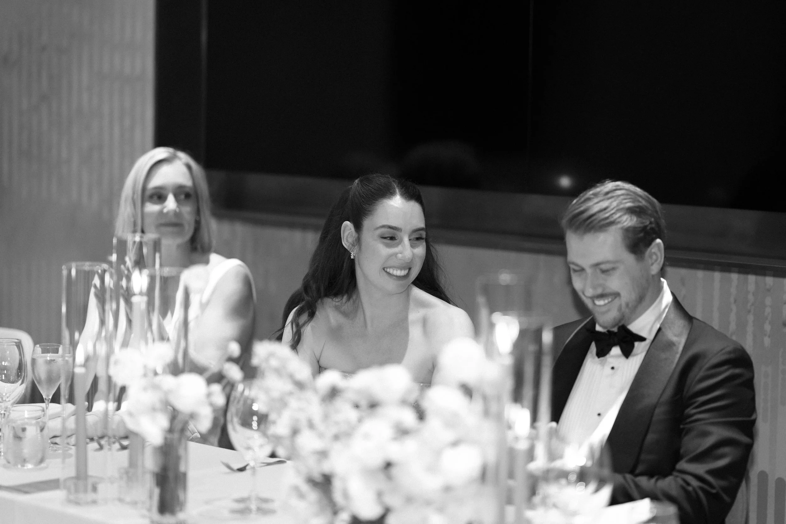Three people dressed in formal attire sitting at a table with floral centerpieces and glassware, smiling and talking at a formal event.