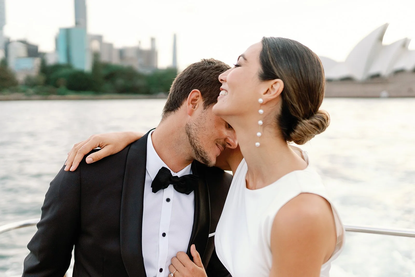 A couple dressed in formal wedding attire sharing an intimate moment by the water with Sydney Opera House and city skyline in the background.