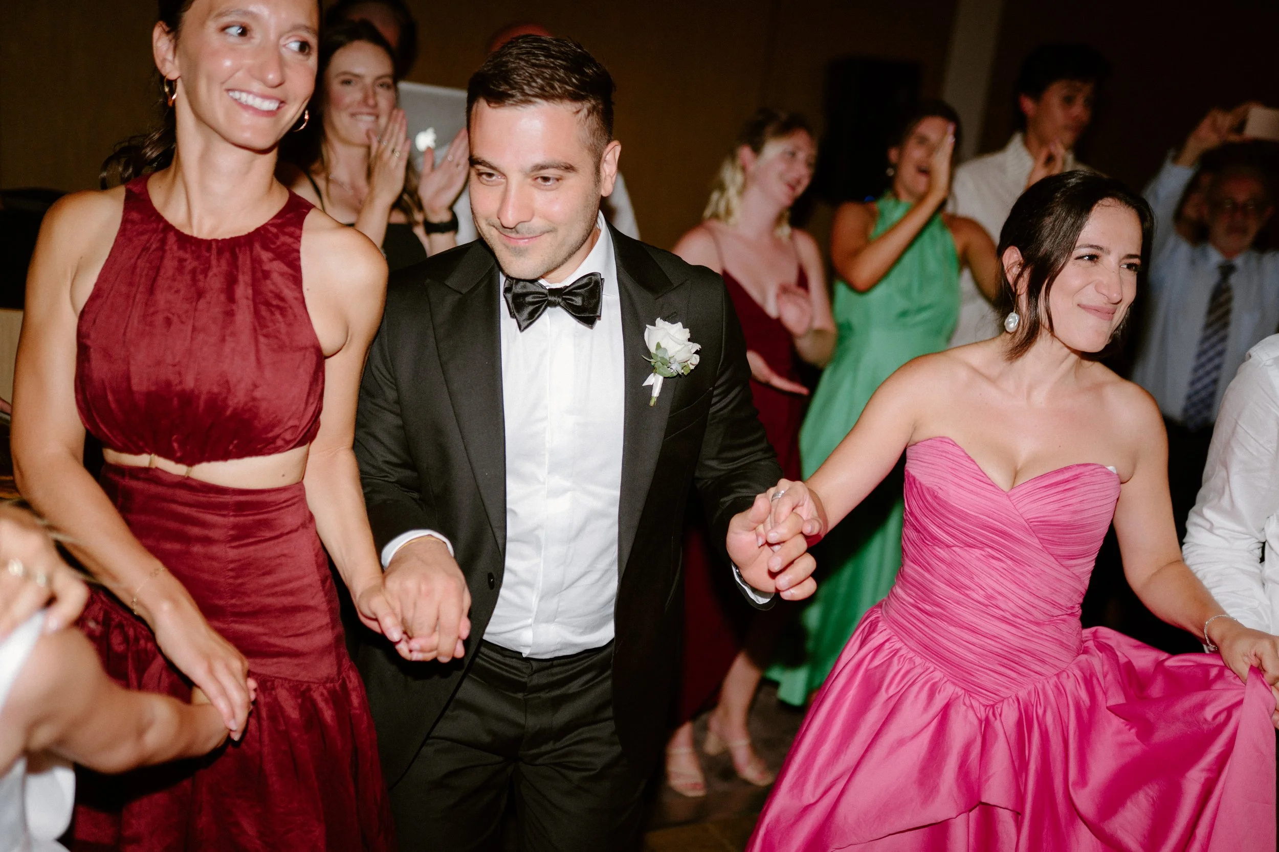 A group of people at a formal celebration or wedding dancing and holding hands, with two women in colorful dresses and a man in a tuxedo.