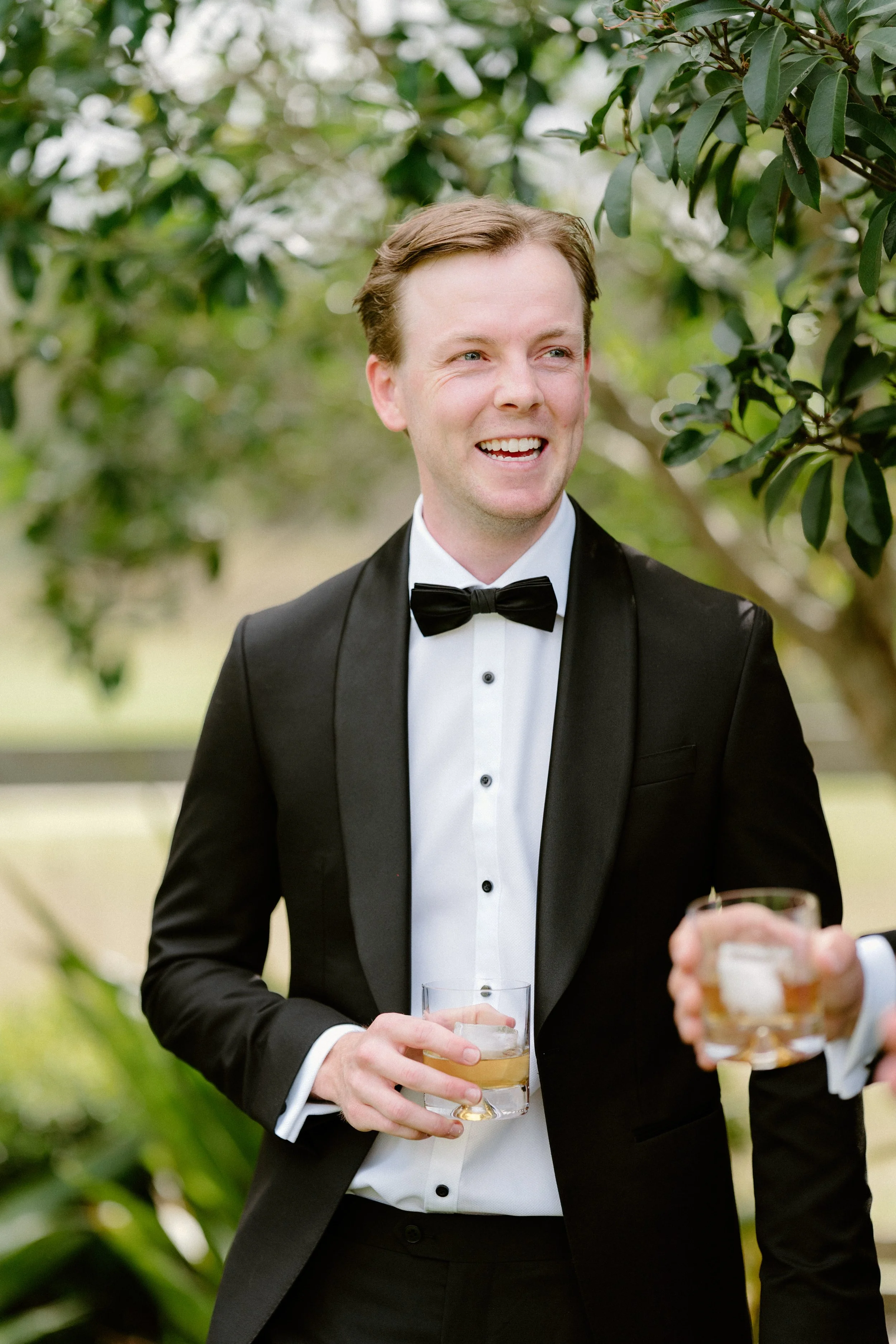 A man dressed in a black tuxedo with a bowtie, smiling and holding a glass of beer at an outdoor event, with greenery in the background.