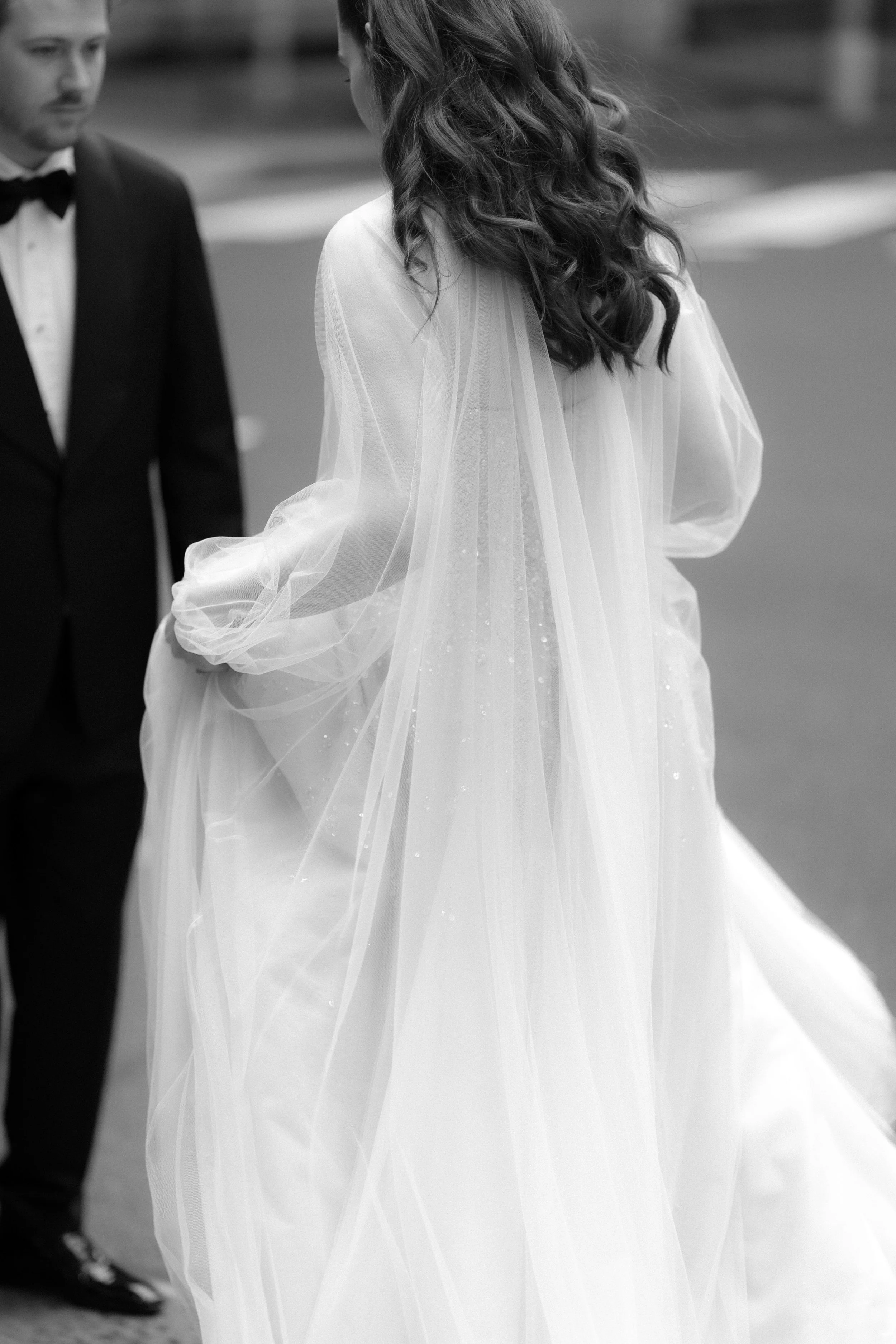 A woman in a wedding dress with a sheer veil, seen from the back, holding up her dress. A man in a tuxedo stands nearby.