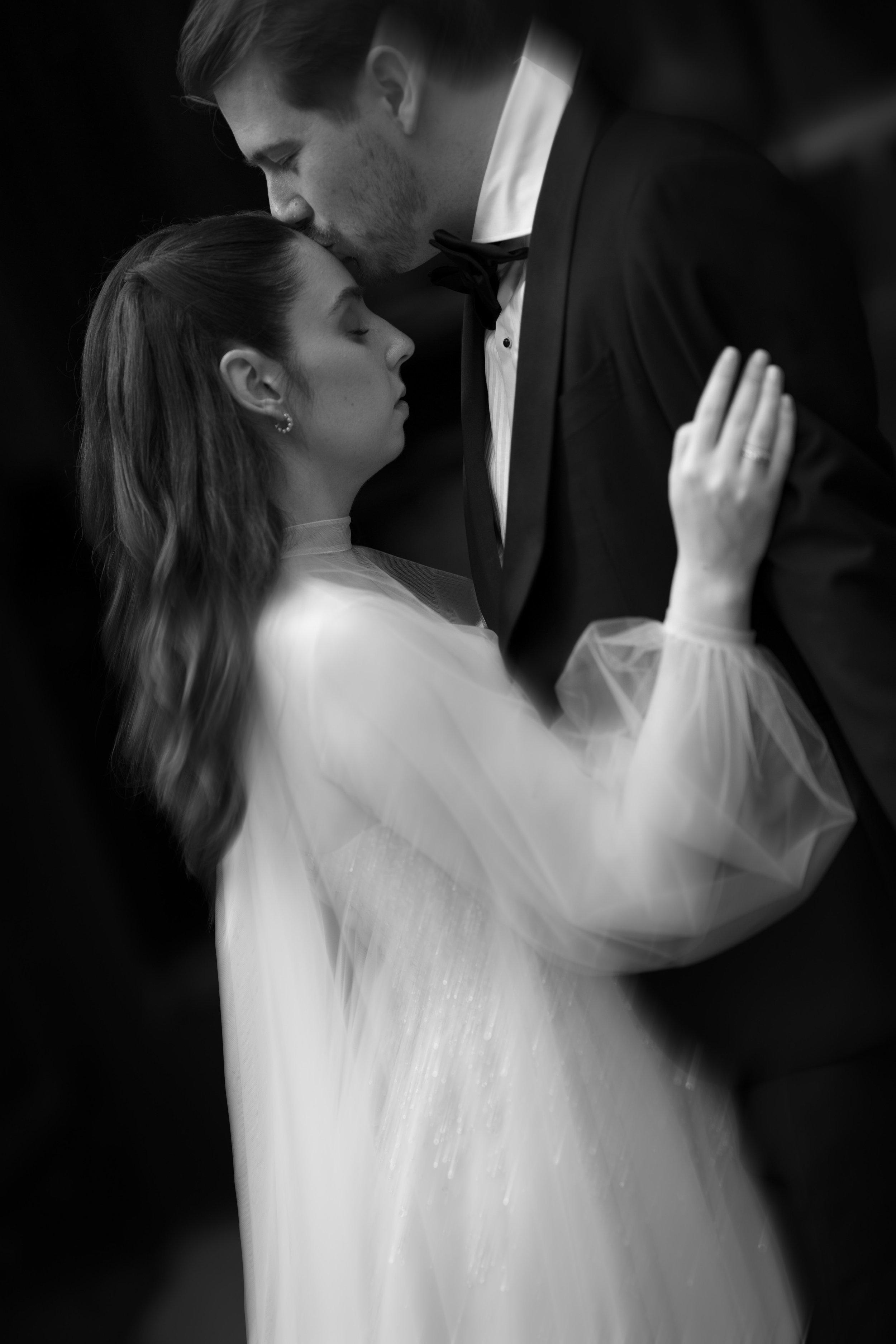 A black and white photo of a couple dancing closely, with the man gently kissing the woman's forehead. The woman has long hair and is wearing a sheer, light-colored dress, while the man is dressed in a tuxedo.