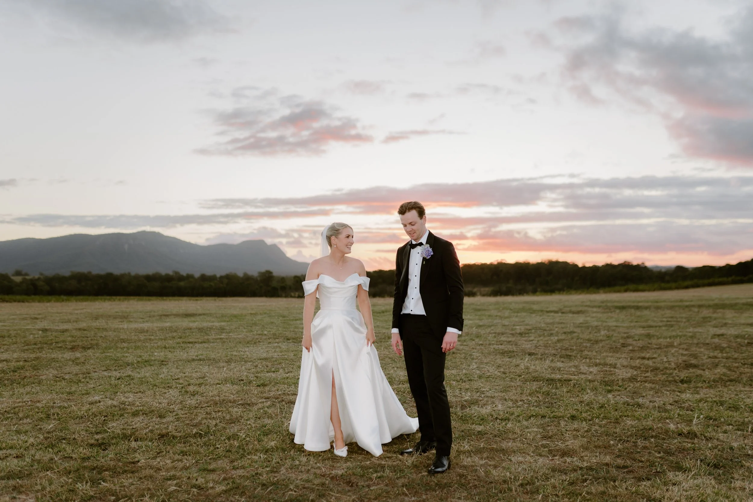 A bride and groom walk together outdoors during sunset in a rural area with mountains in the background. The bride wears a white gown with off-the-shoulder sleeves, and the groom wears a black tuxedo with a bow tie and a purple boutonniere.