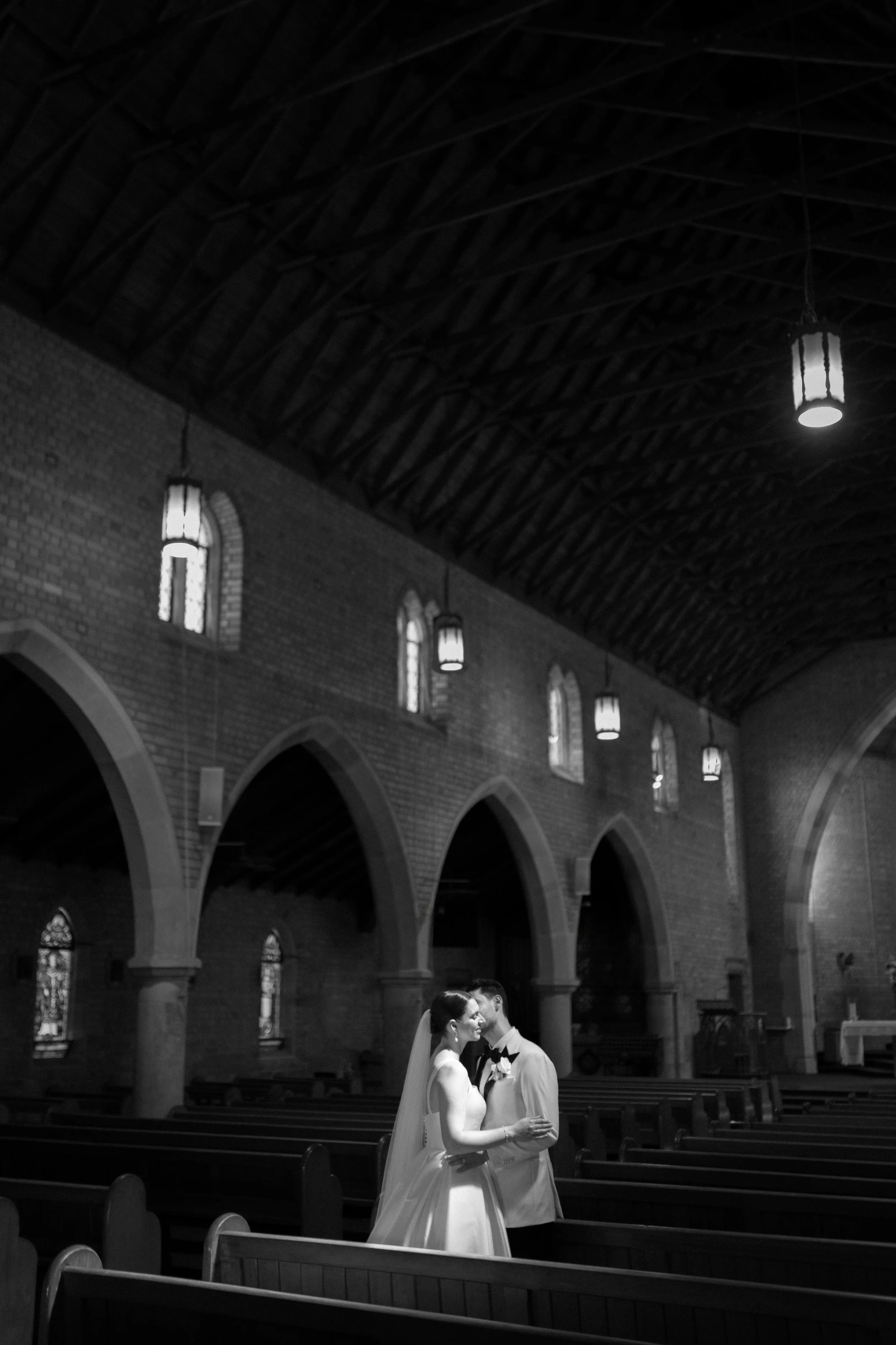 A bride and groom sharing a kiss inside a church with high arched ceilings and hanging lights.