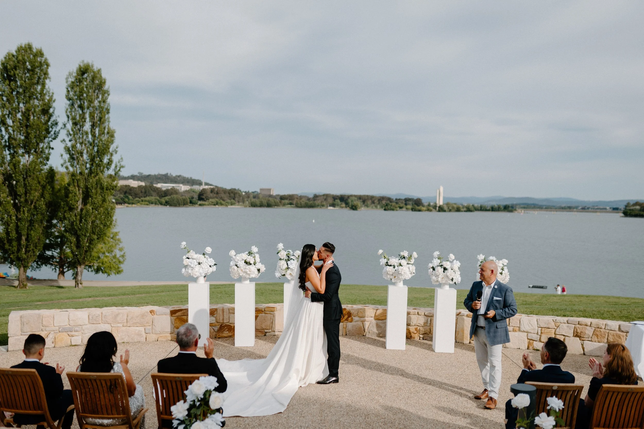Couple getting married outdoors by a lake, with guests seated and a speaker holding a microphone, during daytime with cloudy skies.