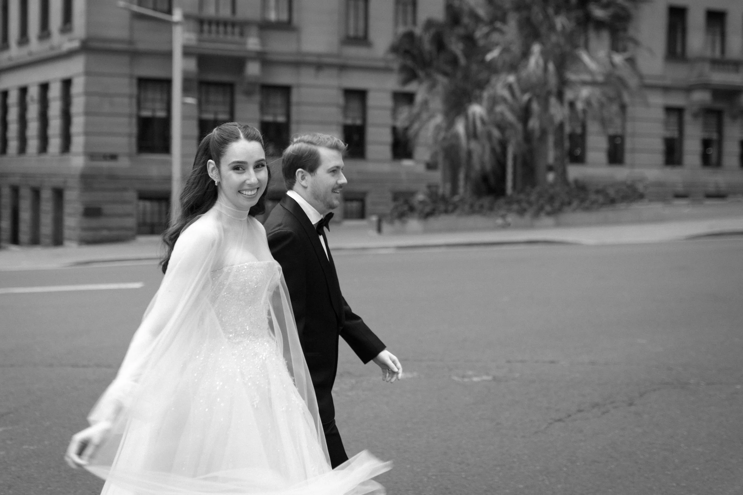 A smiling bride in a wedding dress and a groom in a tuxedo walk on a city street, with a building and palm trees in the background.