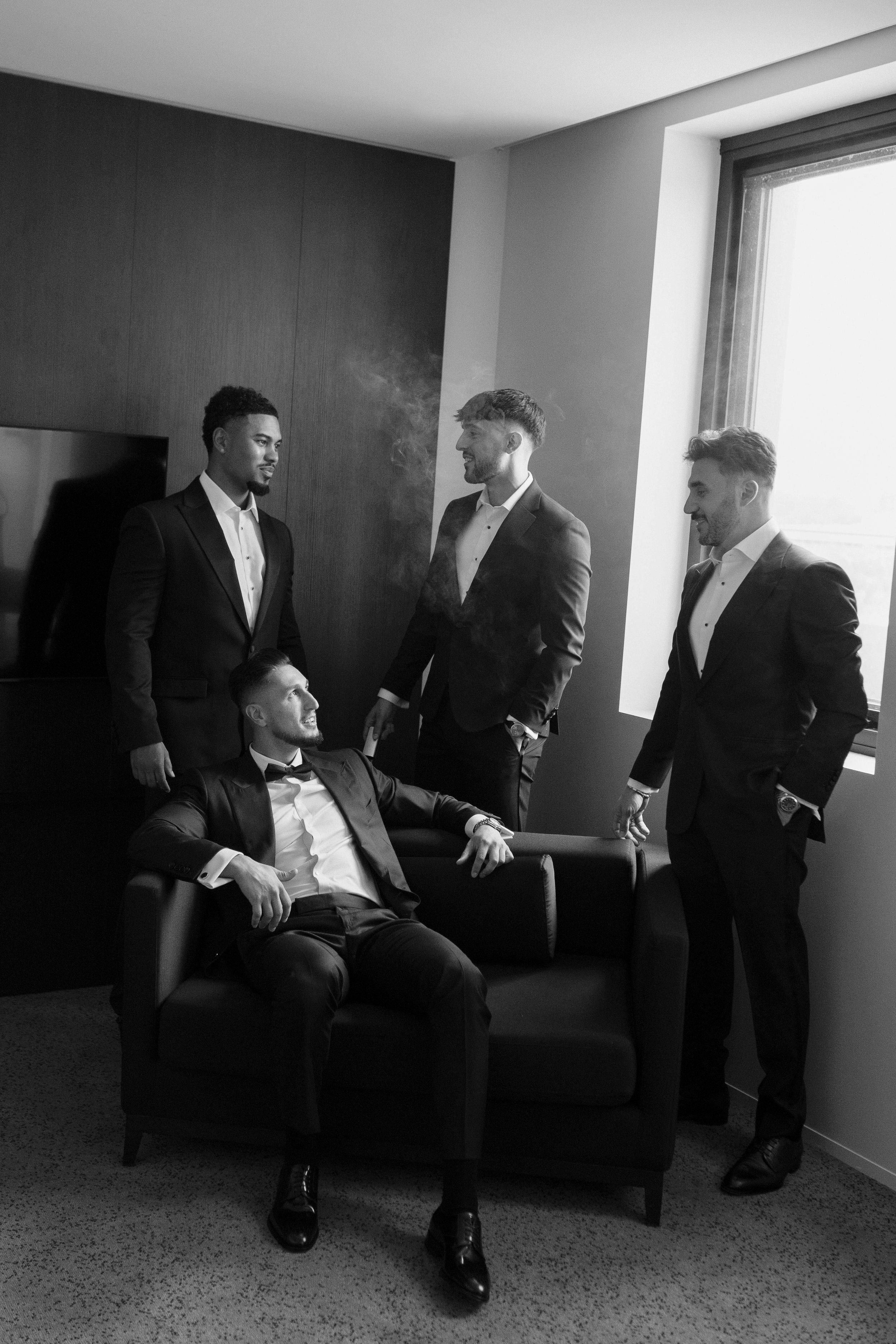 Five young men dressed in tuxedos having a conversation in a modern room with a large window, a television, and a wooden wall panel.
