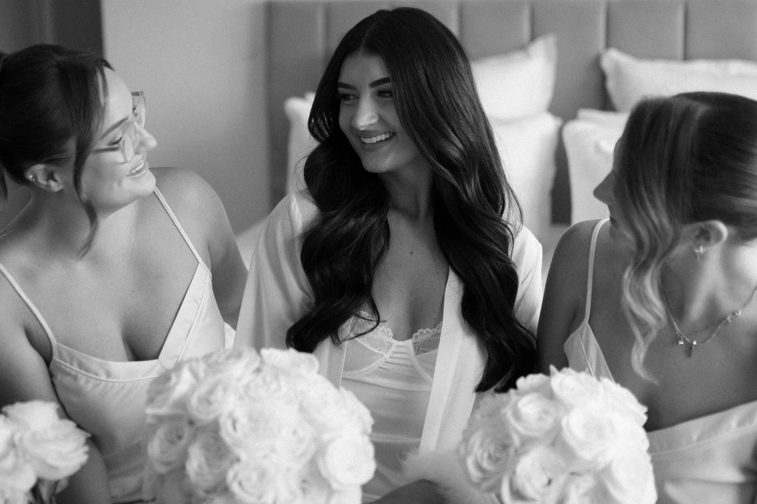 Three women dressed in satin robes, sitting on a bed with white roses, smiling and talking.