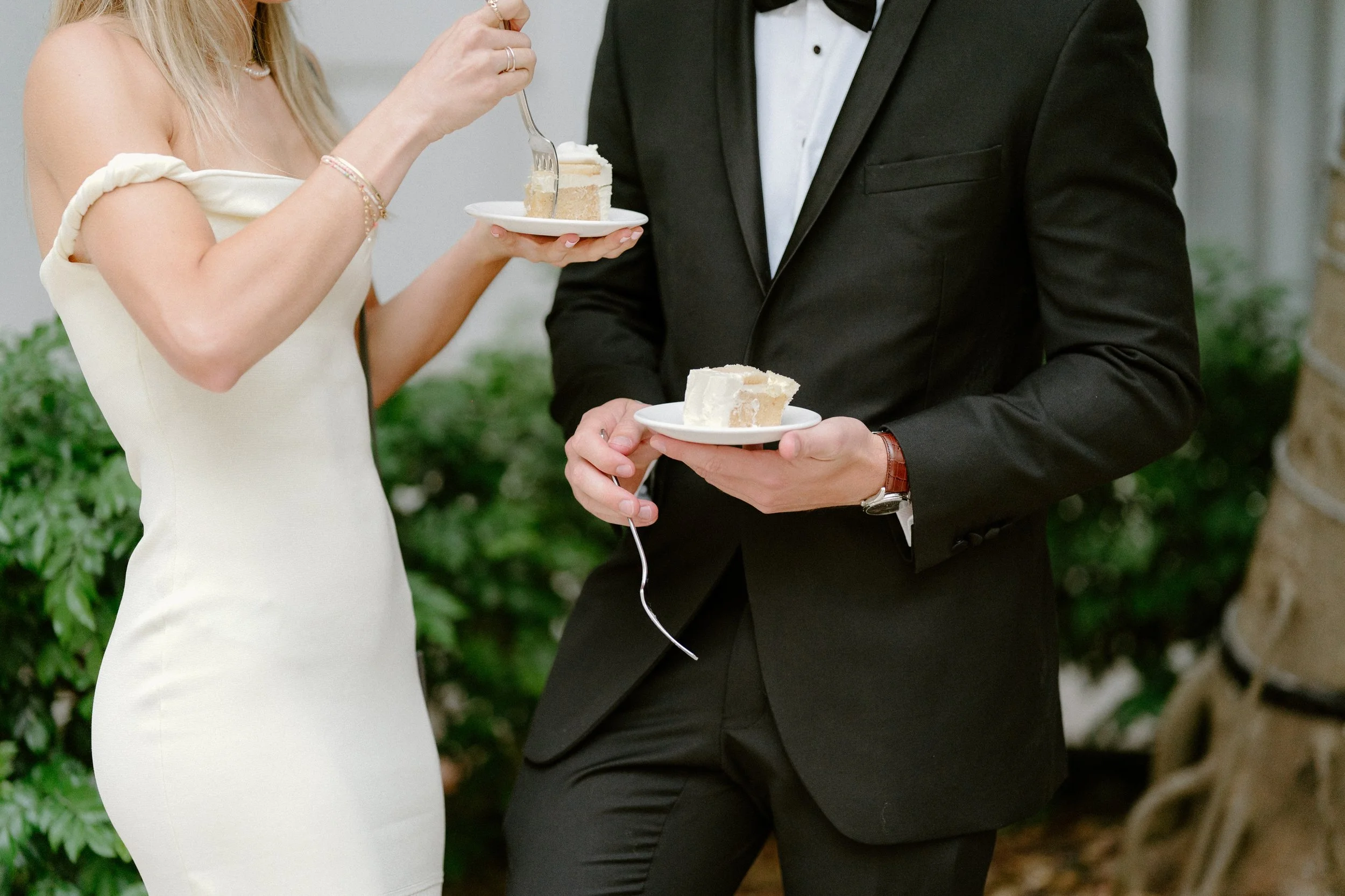 Close-up of a woman and a man at a wedding reception holding small plates with slices of cake, with the woman in a white dress and the man in a black tuxedo.