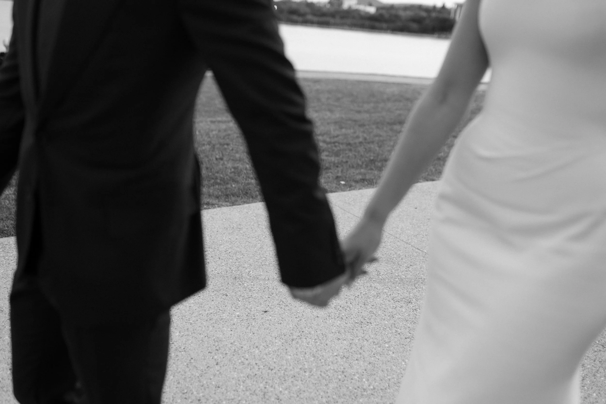 A couple holding hands, the woman in a light dress and the man in a dark suit, walking outdoors near a body of water.