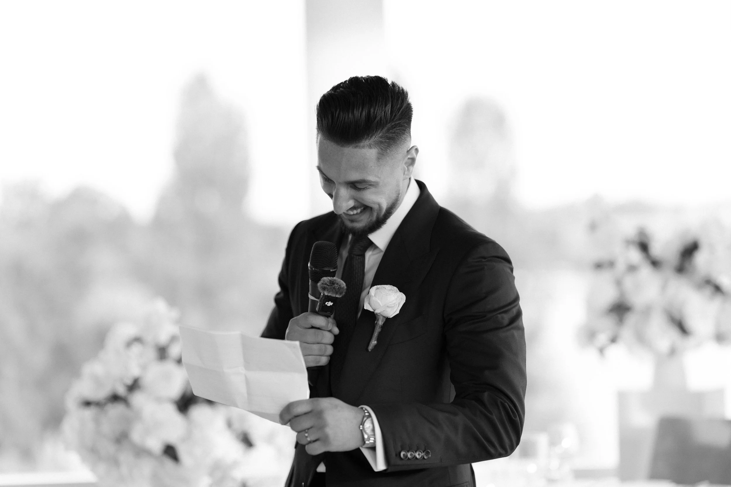 A man in a suit giving a speech at a wedding, holding a paper and a microphone, with a flower boutonniere on his lapel, smiling.