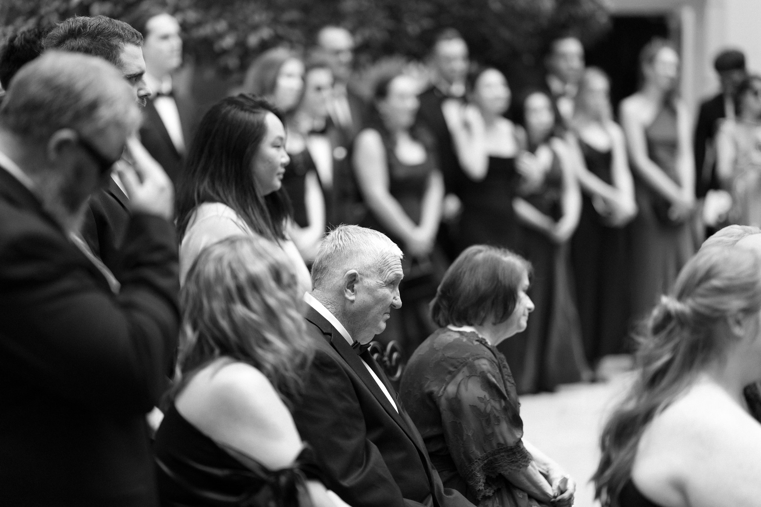 People attending a formal event, some are standing with hands clasped, others are seated and praying, all wearing formal attire, some with face masks.