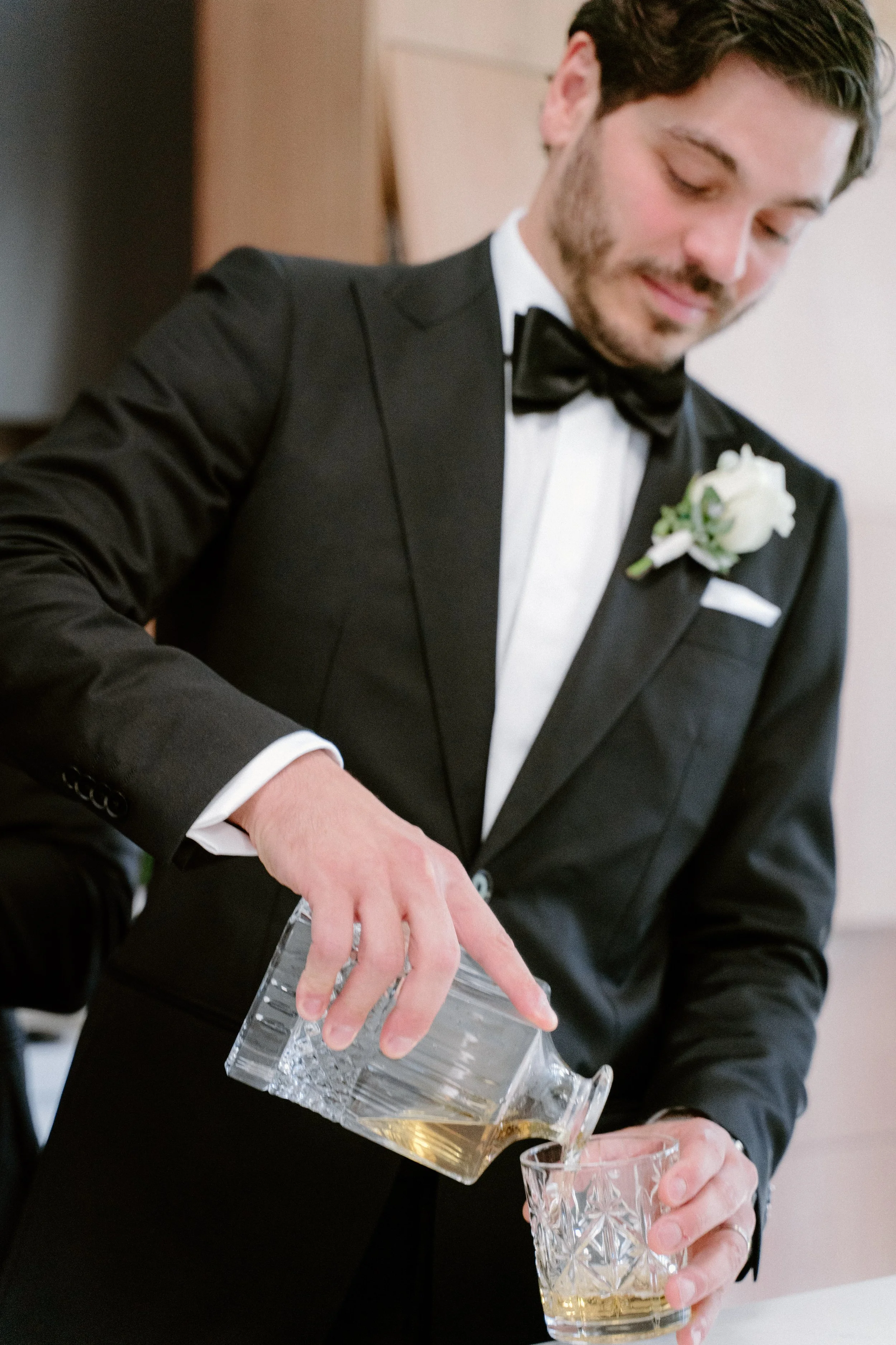A groom in a tuxedo pouring whiskey into a glass at a wedding reception.