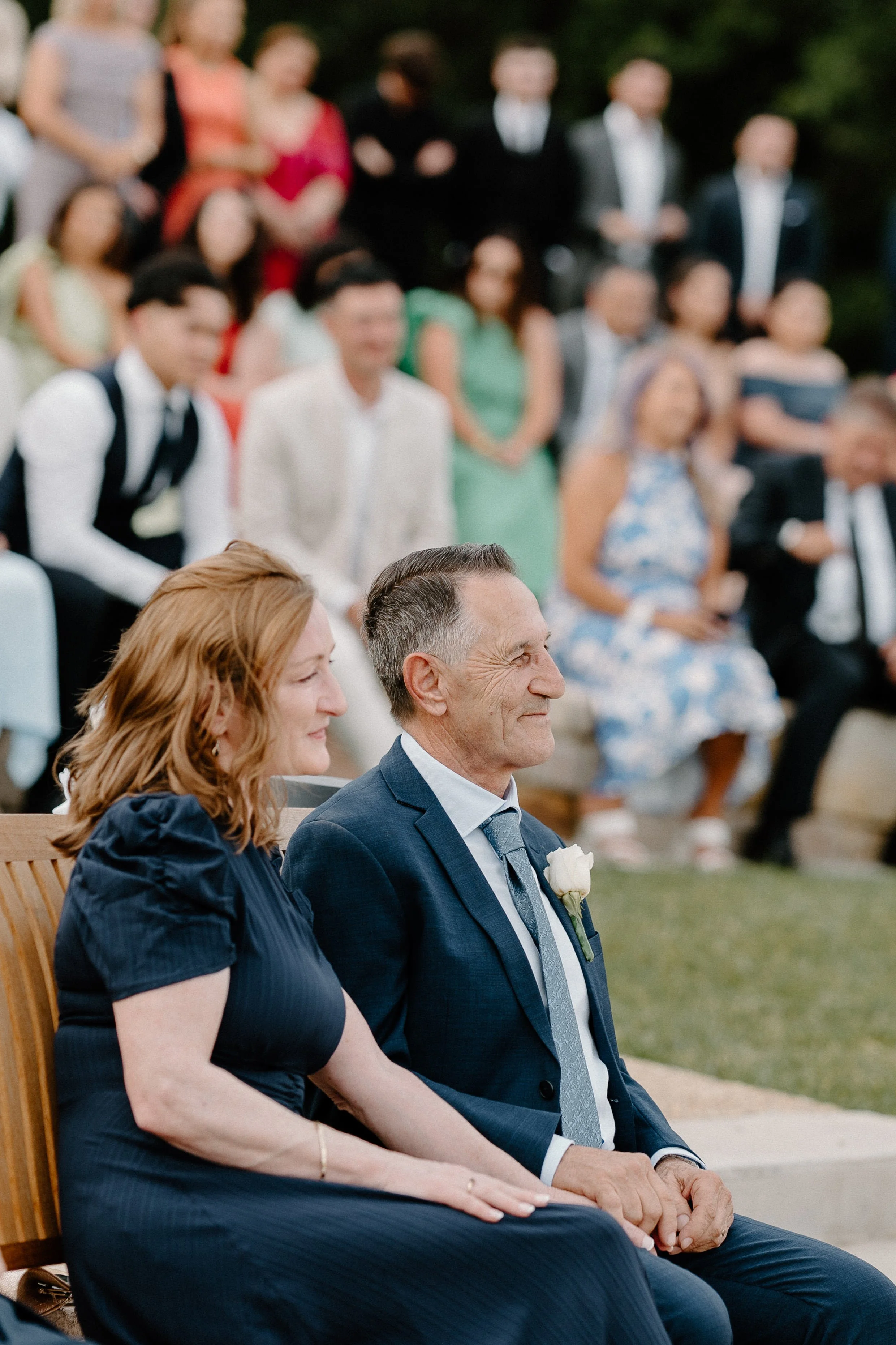 A middle-aged man and woman sitting outdoors during a wedding ceremony, surrounded by guests.