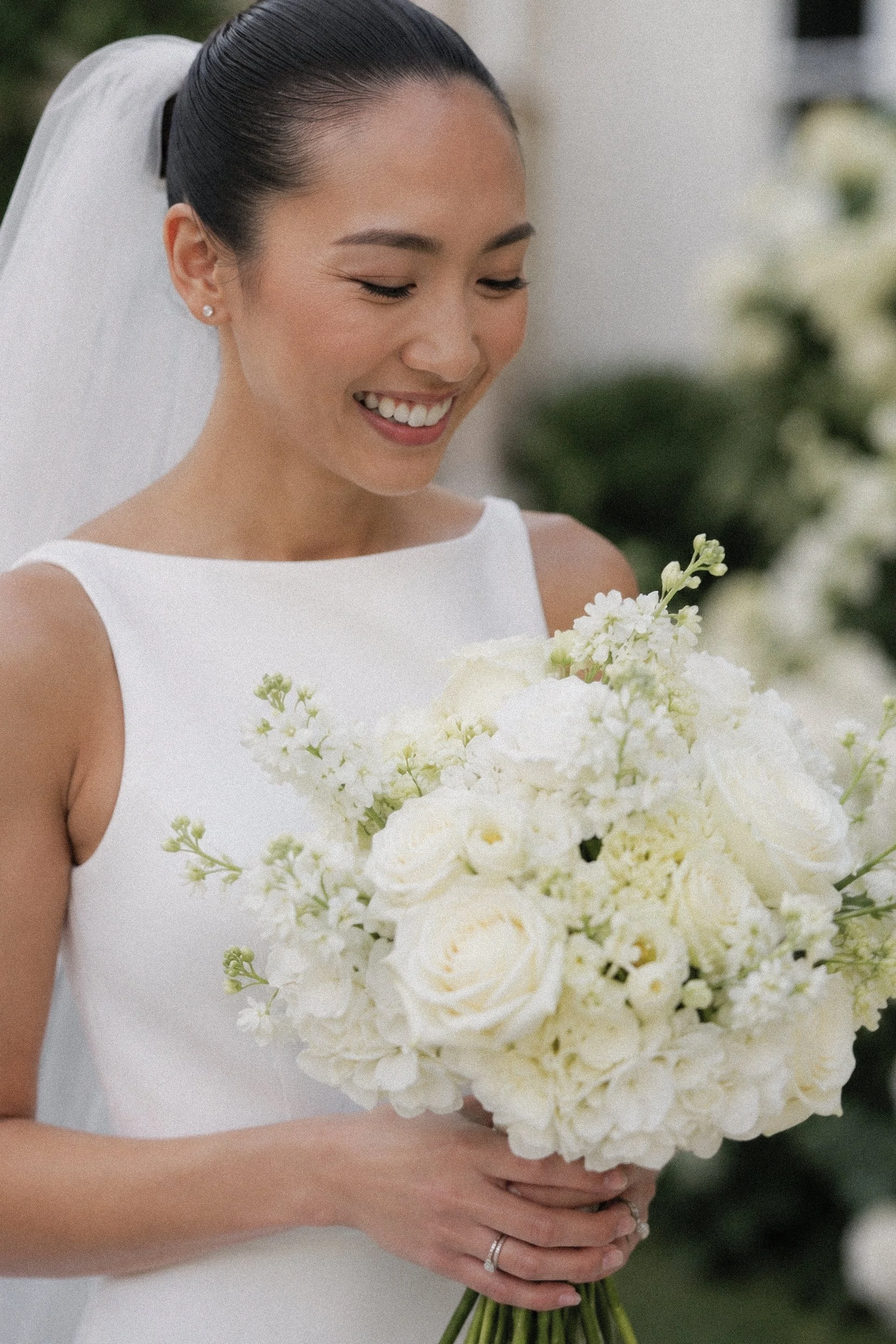 A bride holding a bouquet of white flowers, smiling with eyes closed, wearing a white dress, earrings, and a wedding ring.