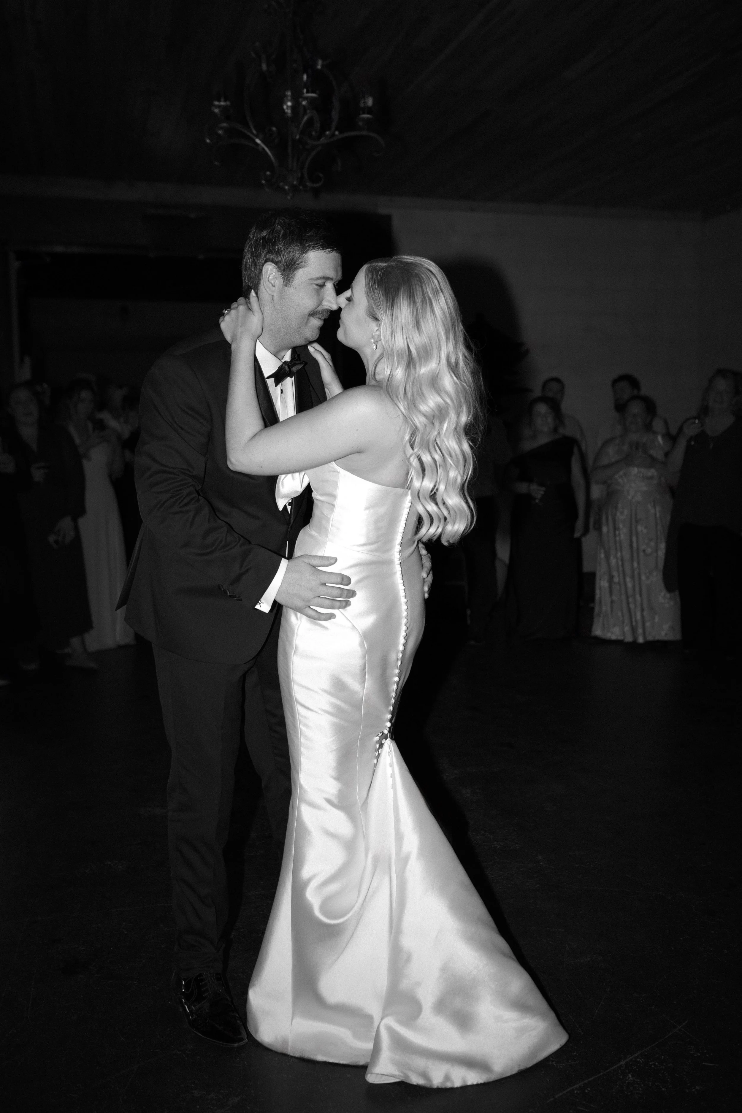 A black and white photo of a bride and groom sharing their first dance at a wedding reception, surrounded by guests.