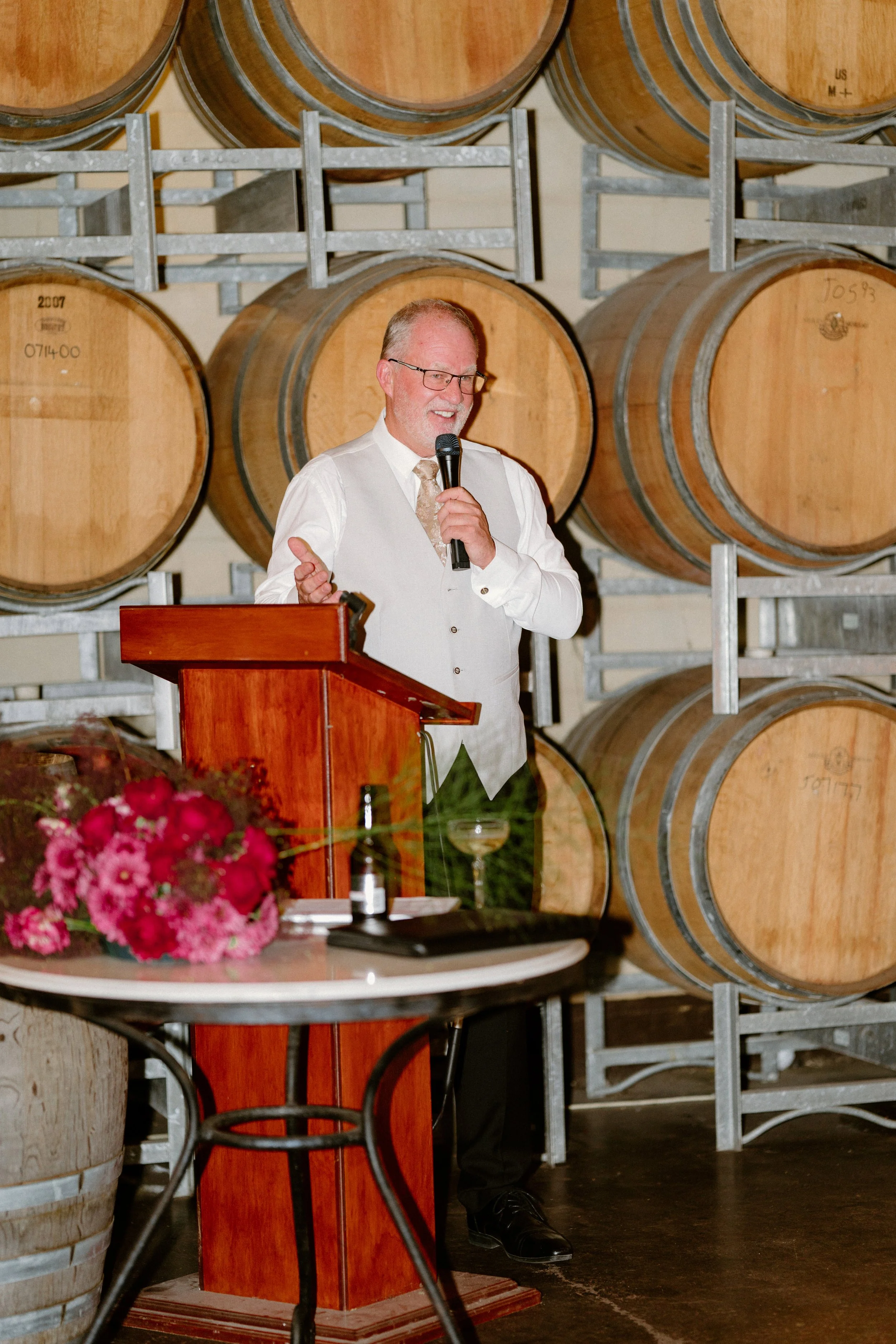 Man in white vest and shirt speaking into a microphone at a wine barrel setting, with flowers and a cocktail on a small table in the foreground.
