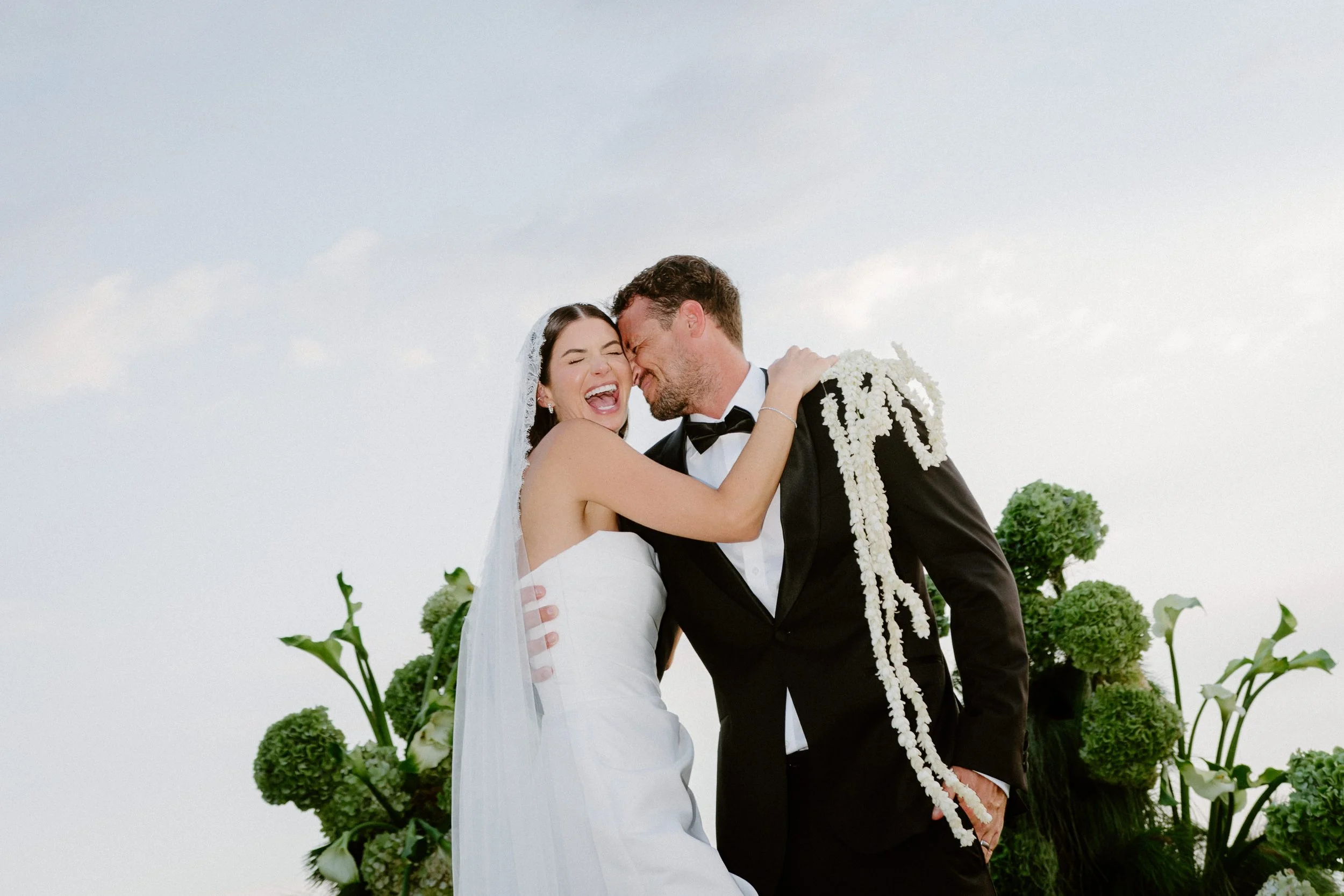 A bride and groom sharing a joyful moment, with the bride laughing and the groom smiling, in an outdoor wedding setting with green plants and a cloudy sky background.