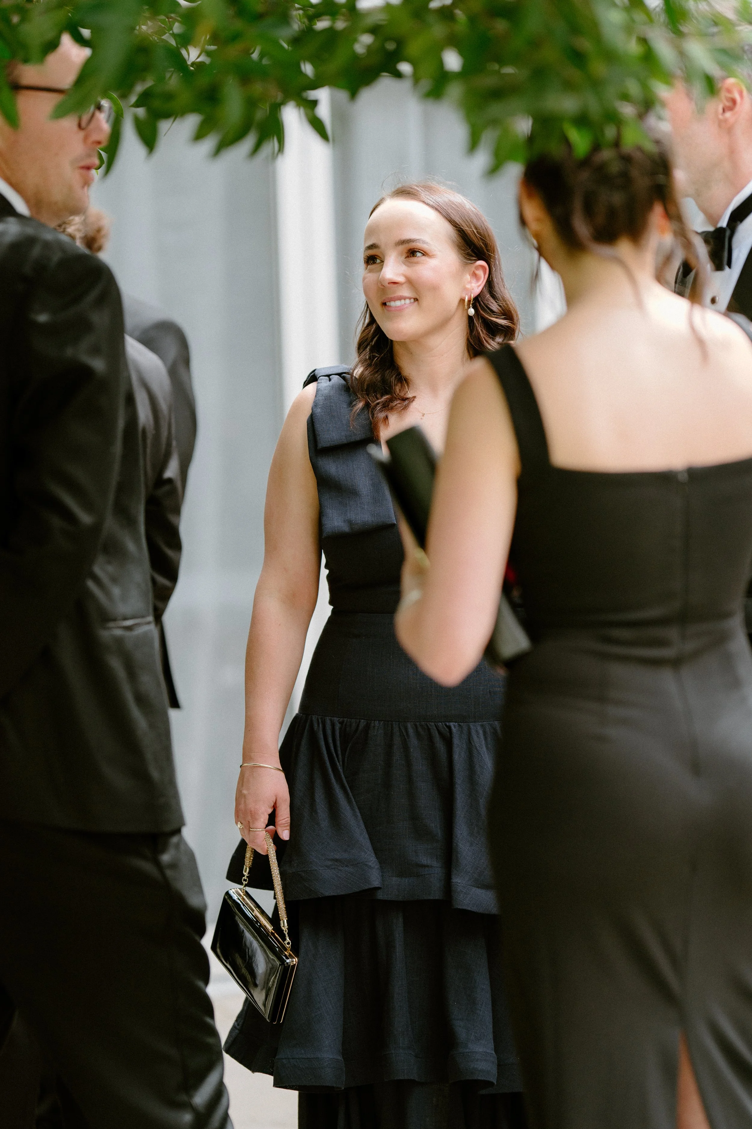 Group of people dressed in formal black attire, possibly at a wedding or formal event, standing outdoors under a tree.