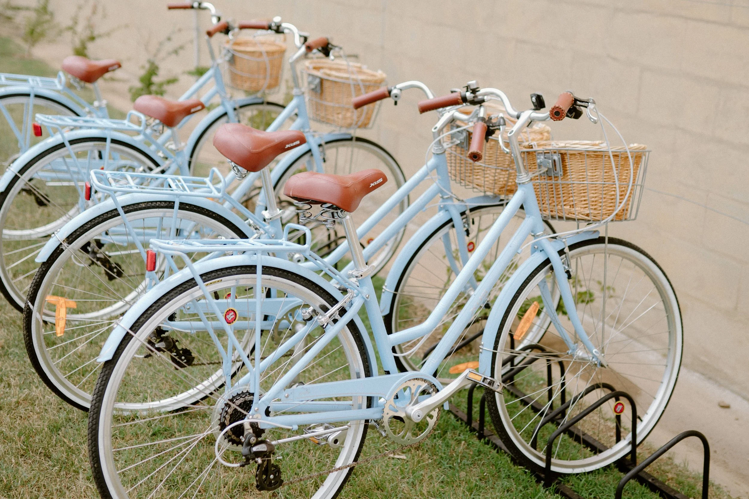 The Sienna, Hunter Valley Wedding Venue, Wedding Photos. Several light blue bicycles with brown seats and baskets are parked in a bike rack on a grassy area near a beige wall.