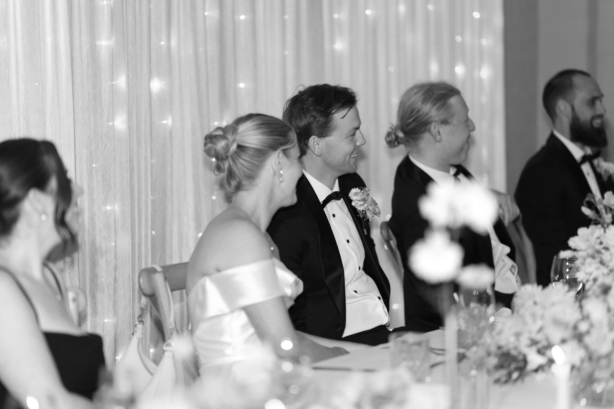 Black and white photo of a wedding reception with a line of people dressed in formal attire, sitting at a table with floral arrangements, and smiling or looking attentive.