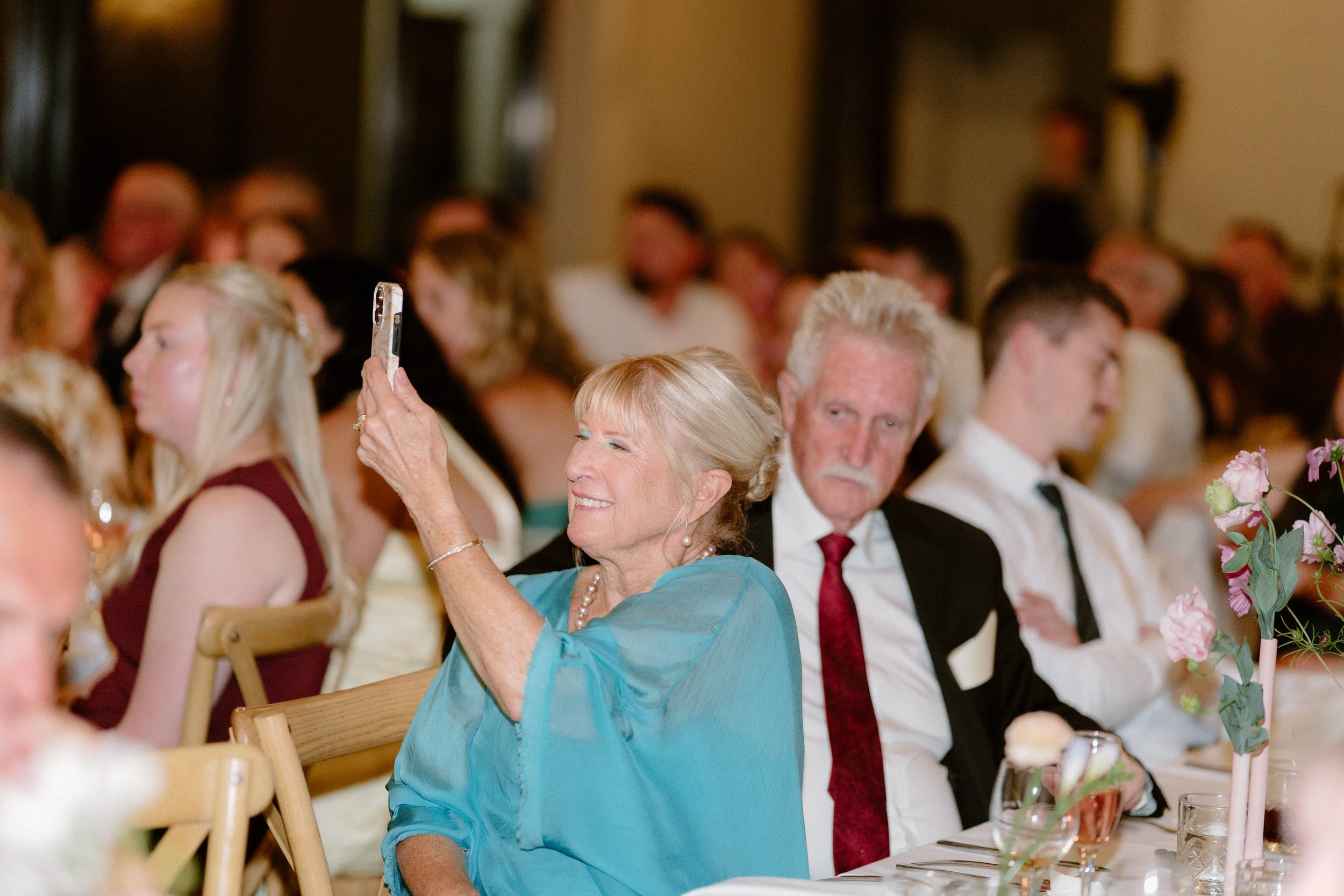 Indoor event with multiple guests seated at tables, including an elderly woman smiling and taking a photo with her smartphone, and a man with gray hair and a woman with blonde hair and a turquoise dress among other attendees.